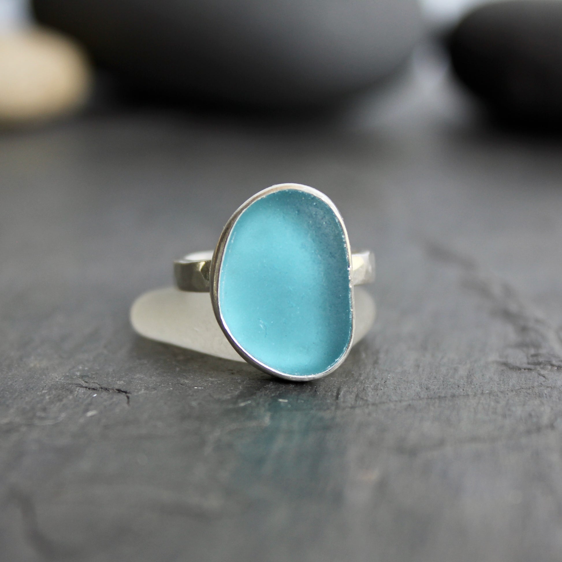 Silver ring with a blue stone on a dark wooden surface.This ring has a large piece of aqua turquoise blue sea glass set in a fine and sterling silver bezel on a sturdy hammered band. Size 8 1/2 Handmade by Barb Macy in Corvallis, Oregon.