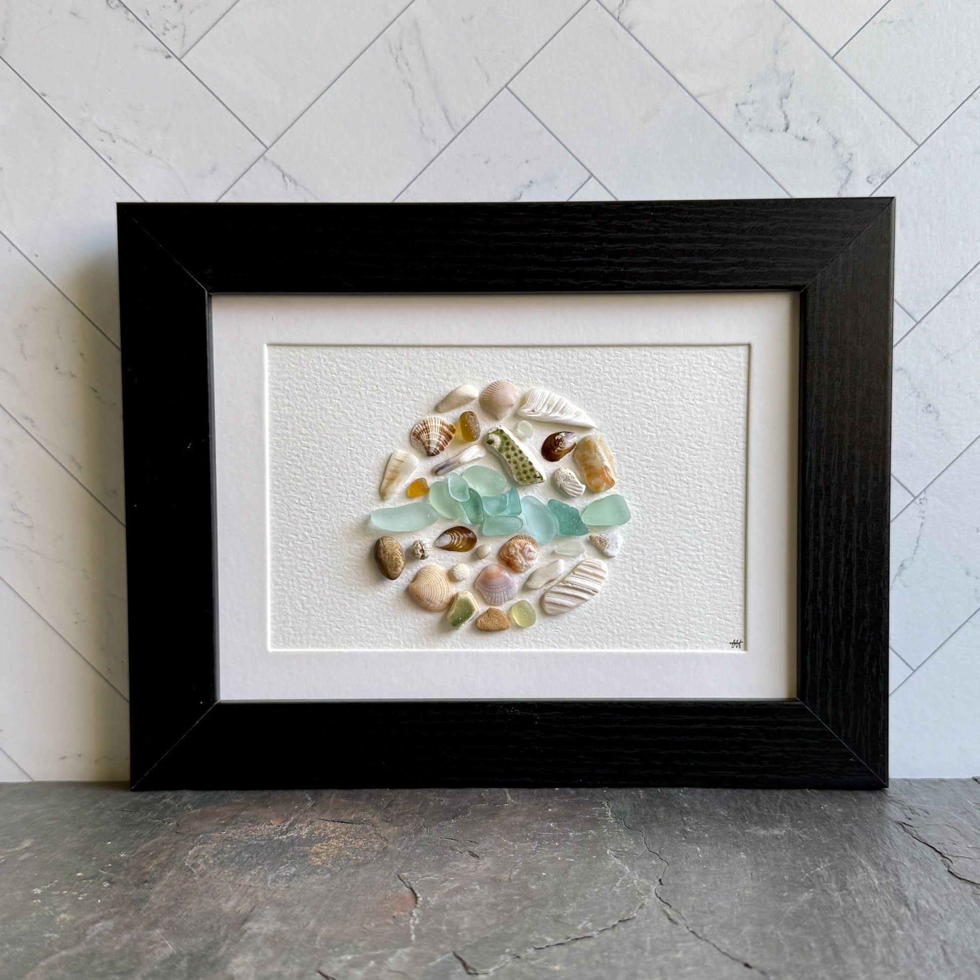 Framed artwork with a circular arrangement of seashells and sea glass on a white background.
