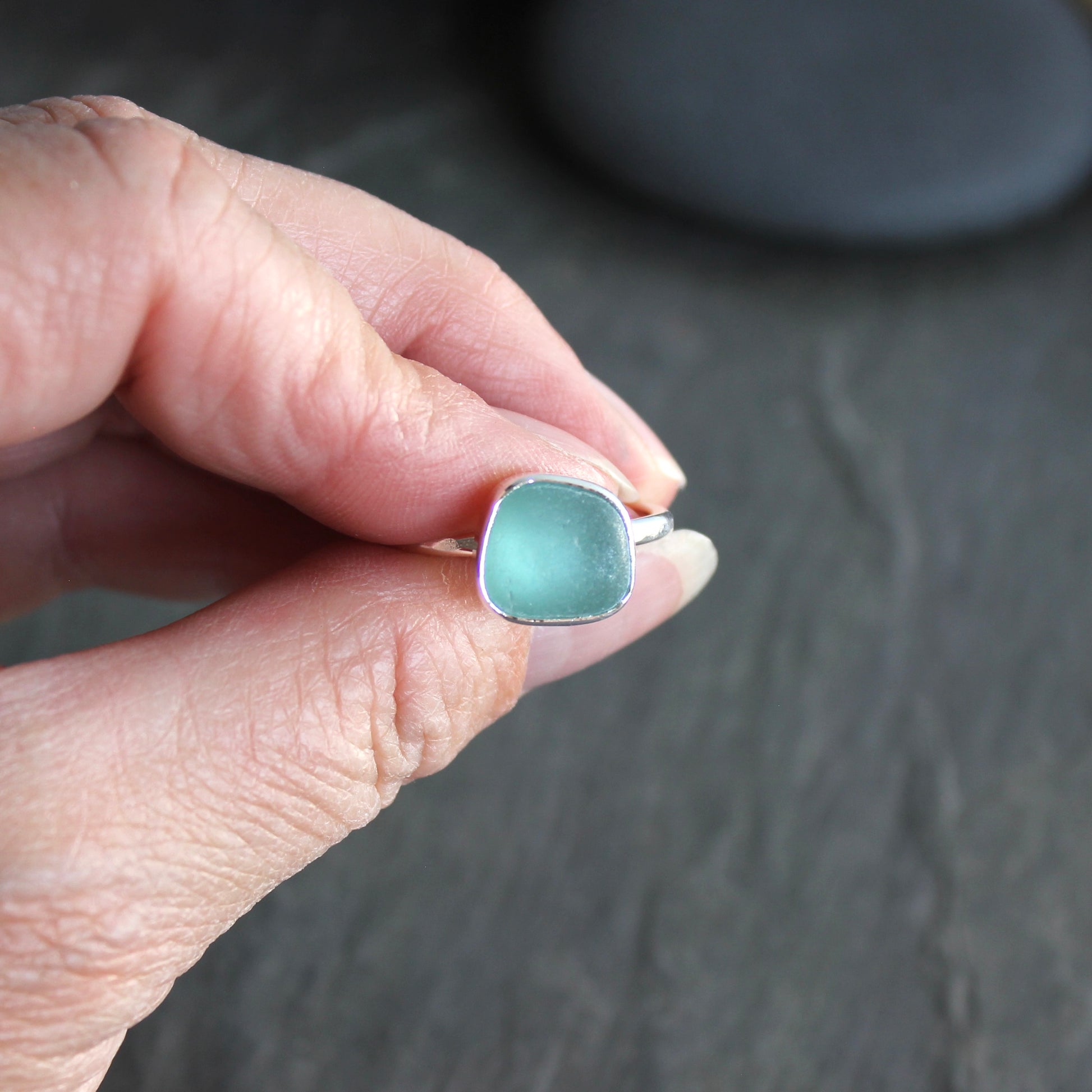 Hand holding a ring with a square, light blue stone on a dark background