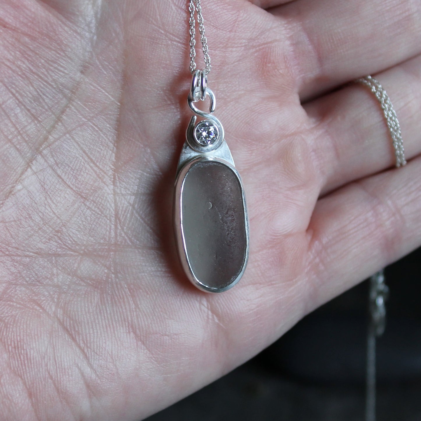 This necklace has a large piece of gray sea glass that is set in a bezel with a simple bail and the back plate cut out so light can shine through the glass. It has a 4mm cubic zirconia to add a little sparkle! It will arrive to you on a sterling silver chain that can be worn at 16, 18 or 20 inches.. All of our sea glass is found and not altered in any way. Handmade by Barb Macy in Corvallis, Oregon. 
