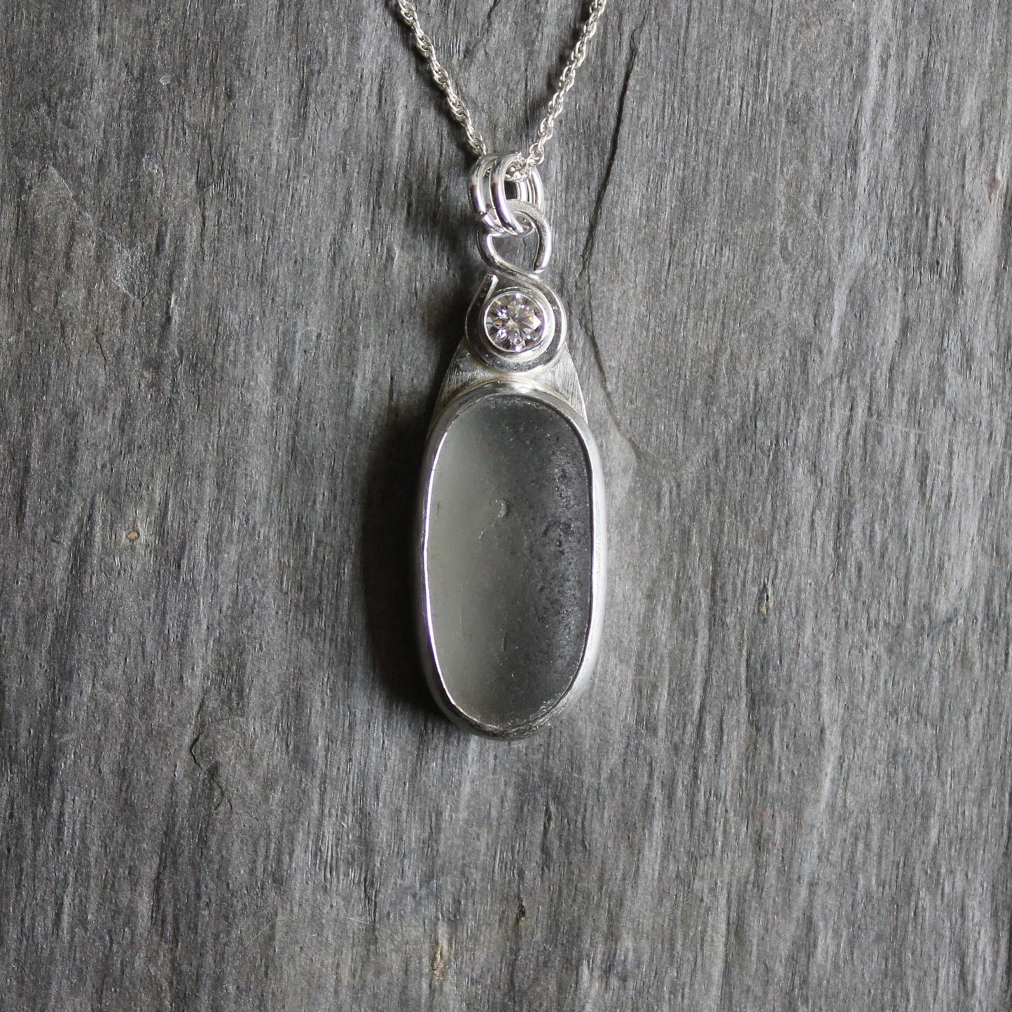 This necklace has a large piece of gray sea glass that is set in a bezel with a simple bail and the back plate cut out so light can shine through the glass. It has a 4mm cubic zirconia to add a little sparkle! It will arrive to you on a sterling silver chain that can be worn at 16, 18 or 20 inches.. All of our sea glass is found and not altered in any way. Handmade by Barb Macy in Corvallis, Oregon. 
