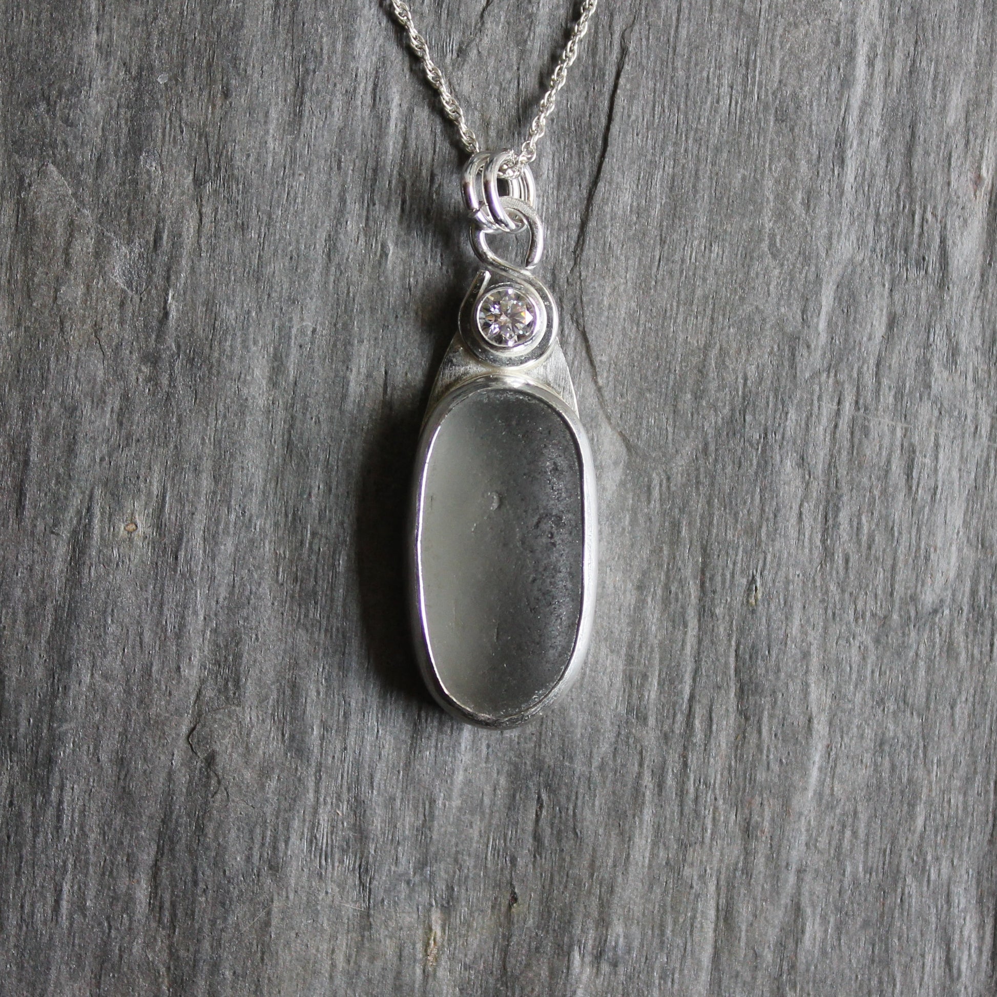 This necklace has a large piece of gray sea glass that is set in a bezel with a simple bail and the back plate cut out so light can shine through the glass. It has a 4mm cubic zirconia to add a little sparkle! It will arrive to you on a sterling silver chain that can be worn at 16, 18 or 20 inches.. All of our sea glass is found and not altered in any way. Handmade by Barb Macy in Corvallis, Oregon. 
