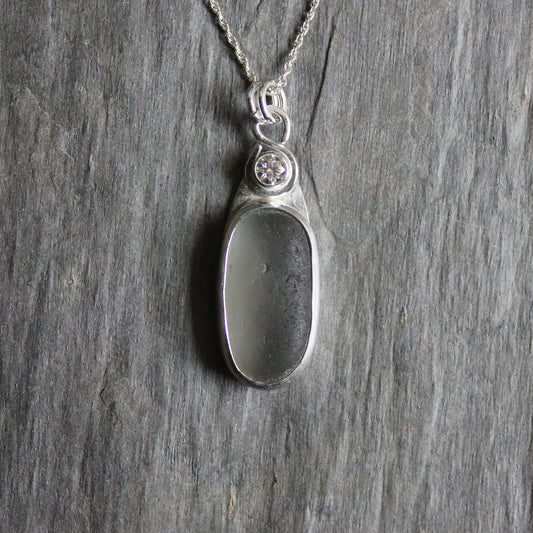 This necklace has a large piece of gray sea glass that is set in a bezel with a simple bail and the back plate cut out so light can shine through the glass. It has a 4mm cubic zirconia to add a little sparkle! It will arrive to you on a sterling silver chain that can be worn at 16, 18 or 20 inches.. All of our sea glass is found and not altered in any way. Handmade by Barb Macy in Corvallis, Oregon. 
