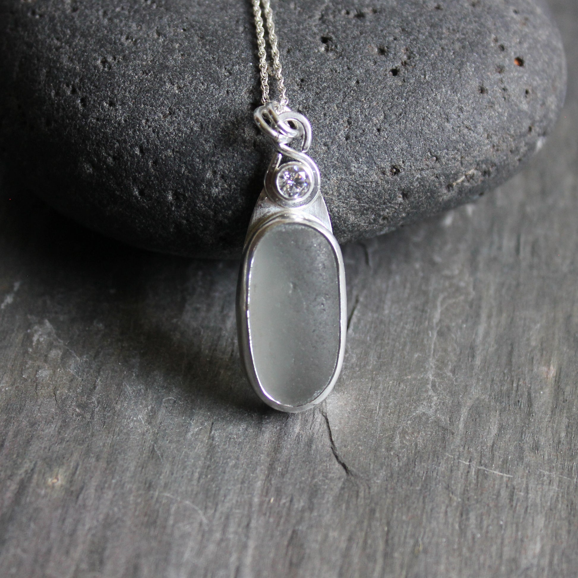 This necklace has a large piece of gray sea glass that is set in a bezel with a simple bail and the back plate cut out so light can shine through the glass. It has a 4mm cubic zirconia to add a little sparkle! It will arrive to you on a sterling silver chain that can be worn at 16, 18 or 20 inches.. All of our sea glass is found and not altered in any way. Handmade by Barb Macy in Corvallis, Oregon. 
