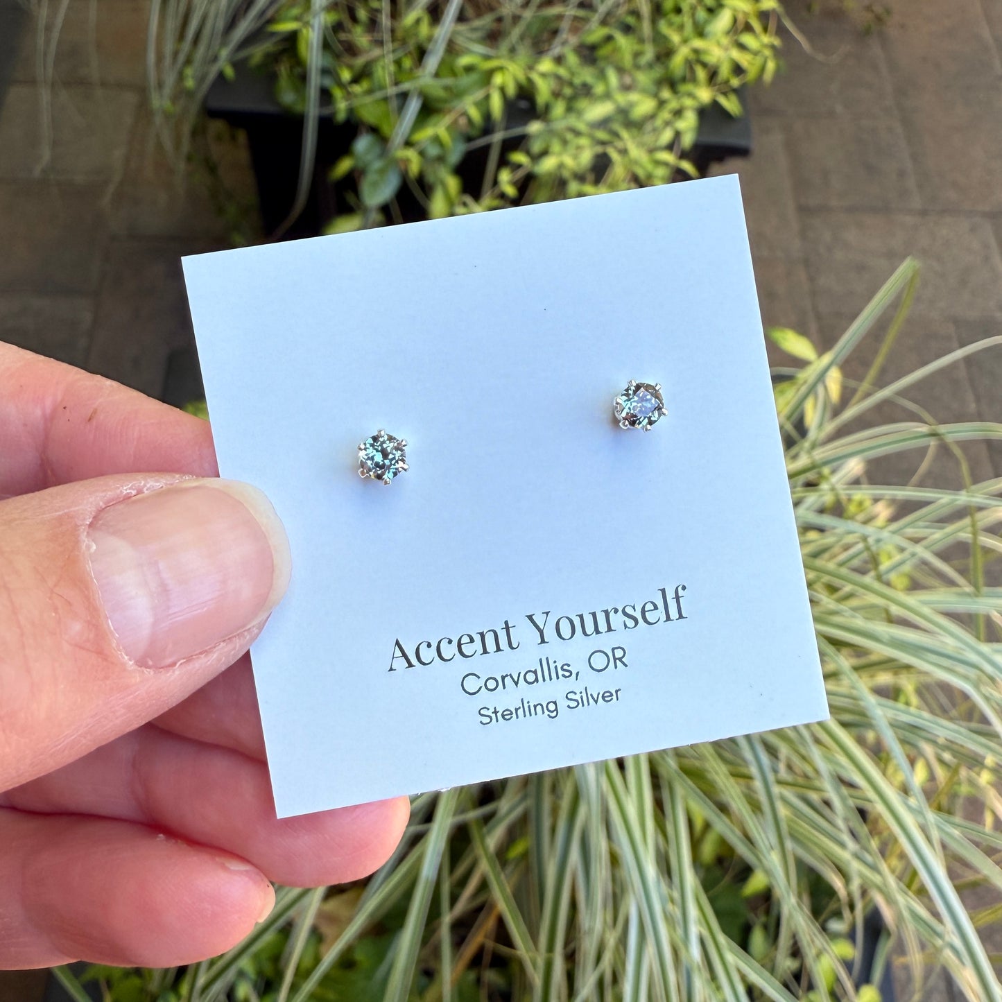 Silver stud earrings on a 'Accent Yourself' card held by a hand with plants in the background