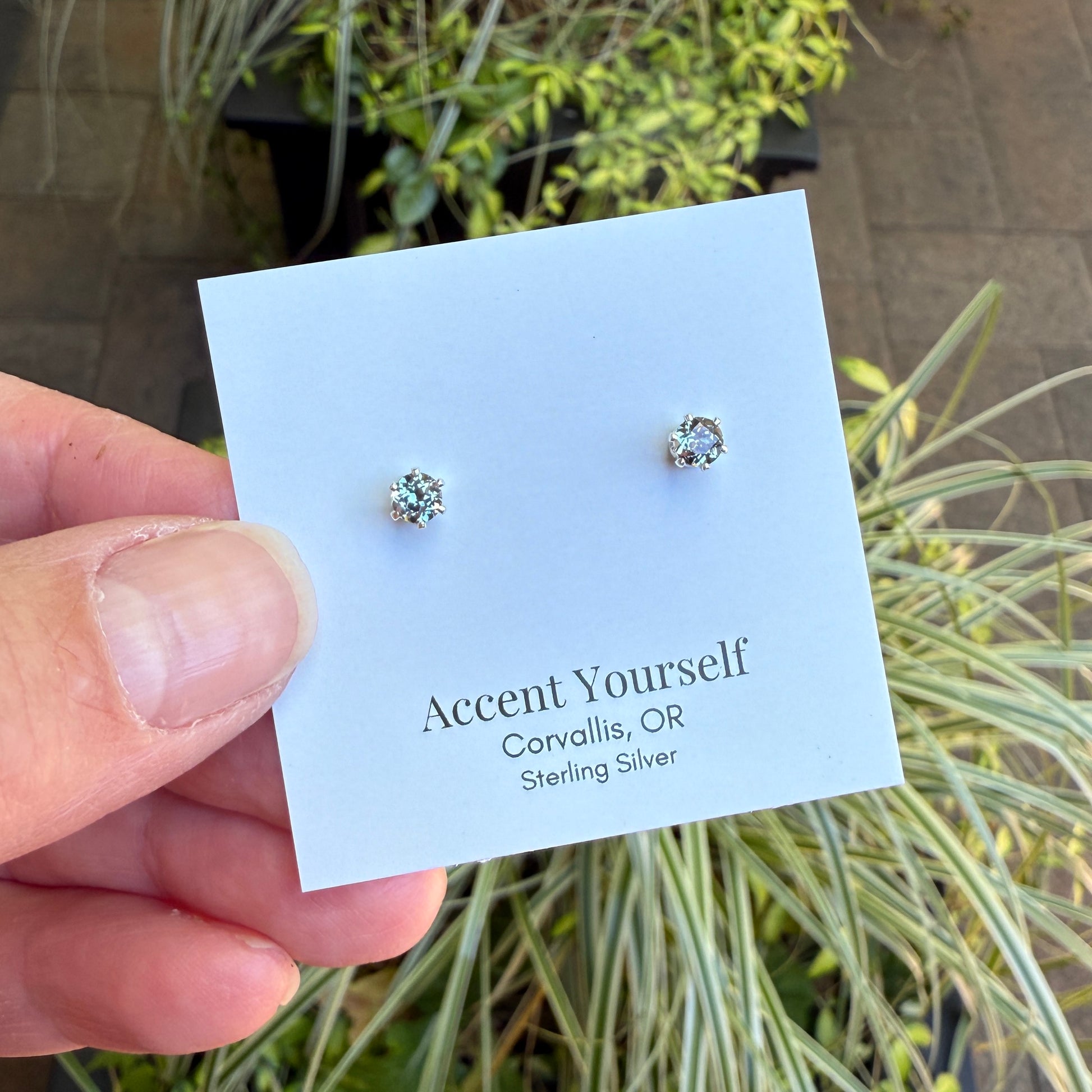 Silver stud earrings on a 'Accent Yourself' card held by a hand with plants in the background