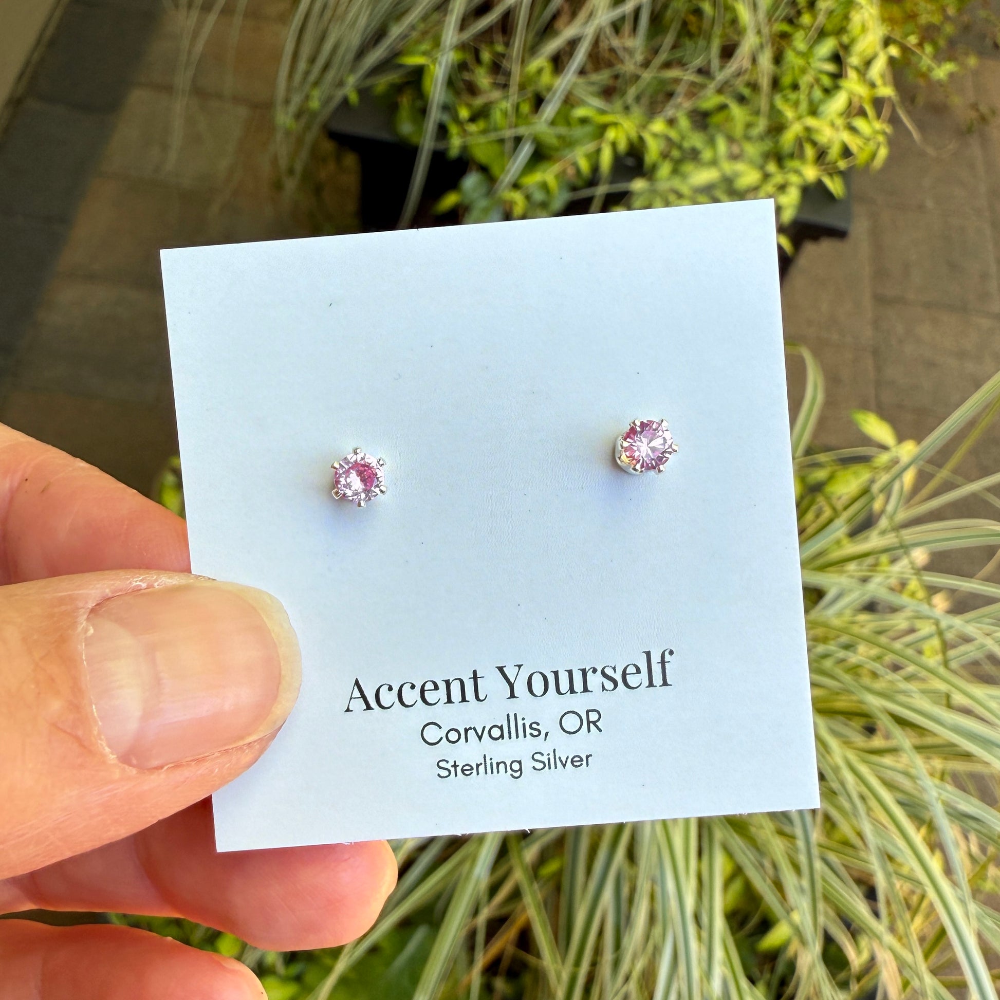 Pink gemstone earrings on a card held by a hand with plants in the background