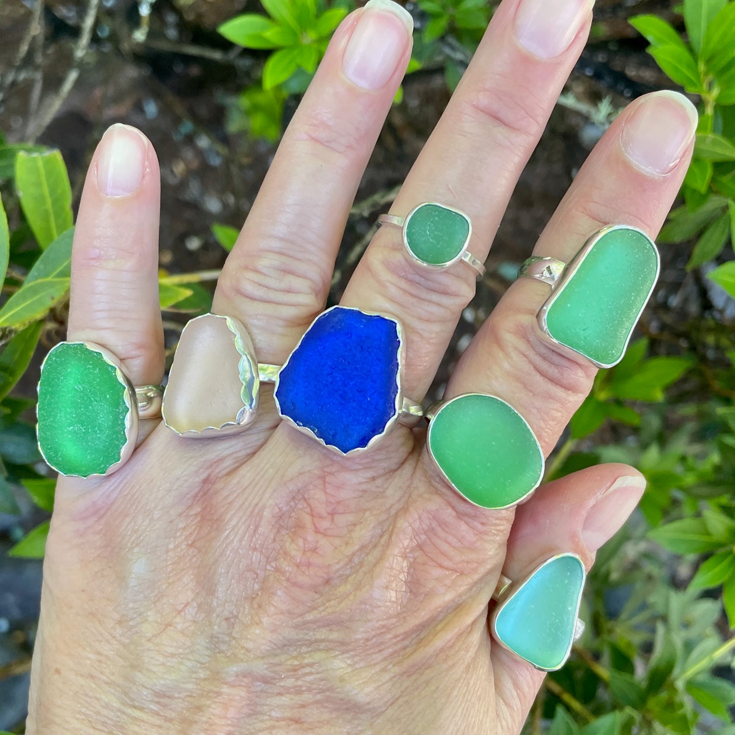 Hand wearing multiple green and blue sea glass ring designs against a natural background