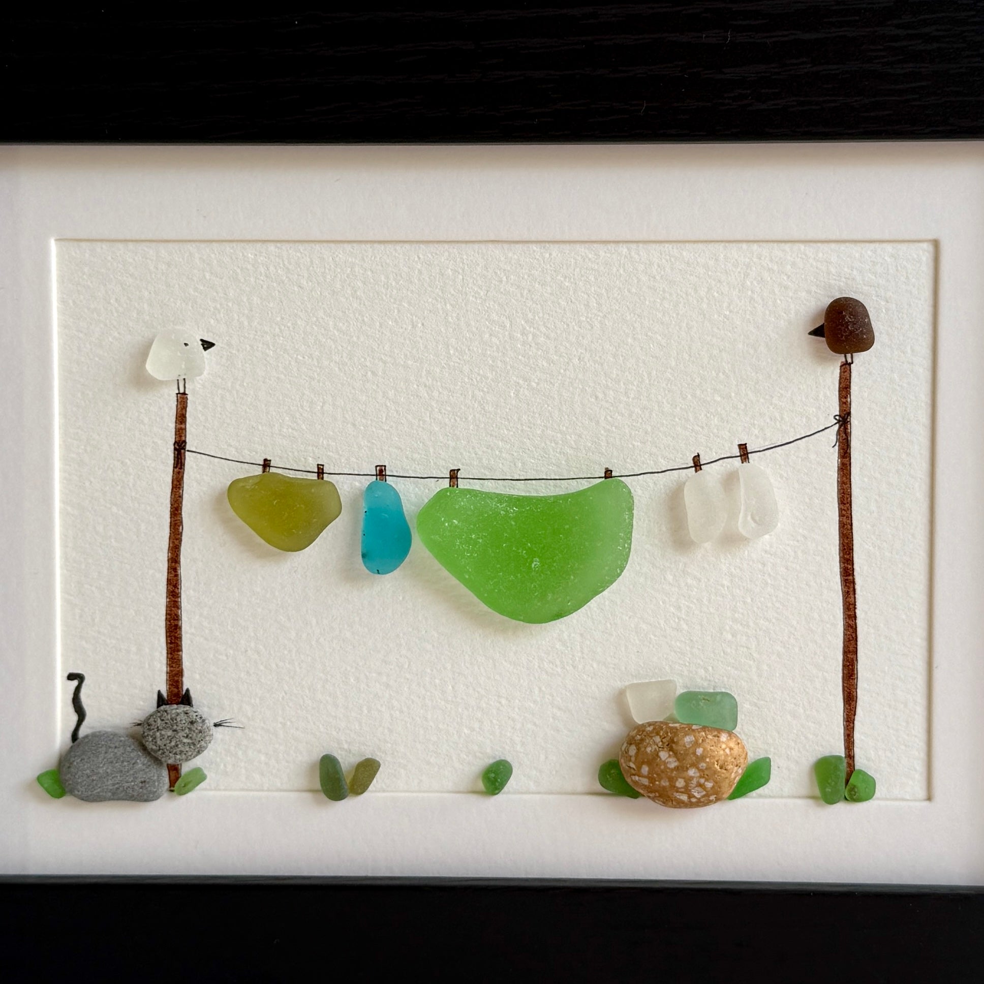 Artistic display of sea glass and stones on a string with wooden sticks and small animals, framed.