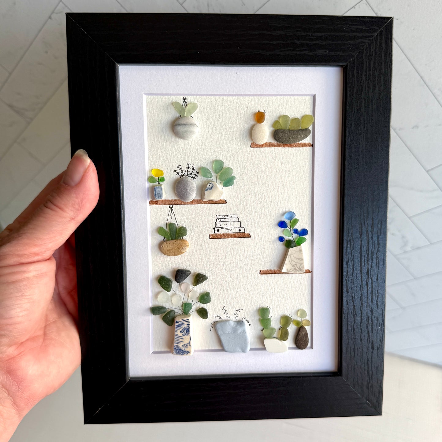 Framed artwork of plants on shelves held by a hand
