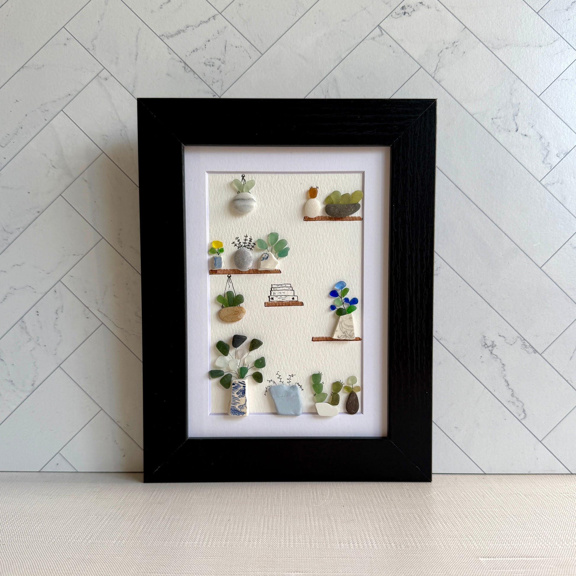 Framed artwork of small plants and objects on shelves against a marble wall.