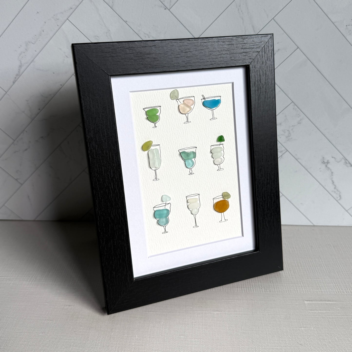 Framed artwork of hand-drawn cocktail glasses filled with colorful sea glass on a white background.