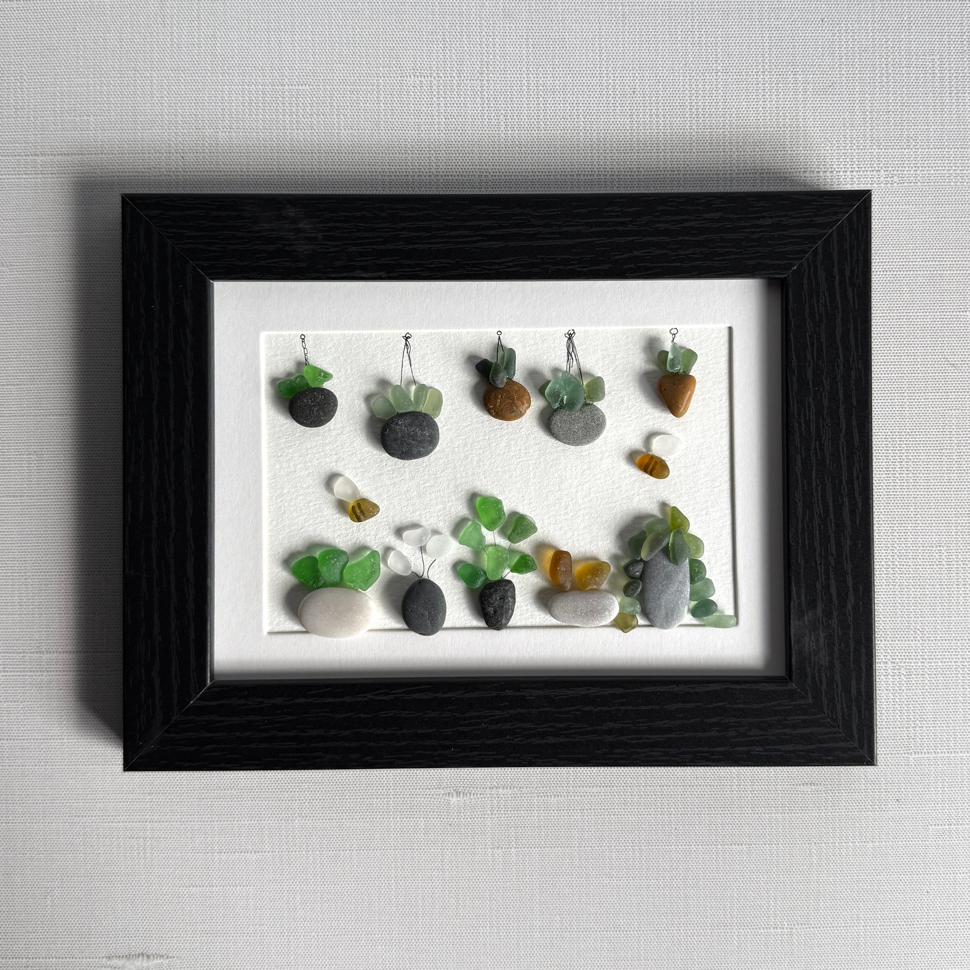 Framed artwork of small plants made from stones and sea glass on a white background