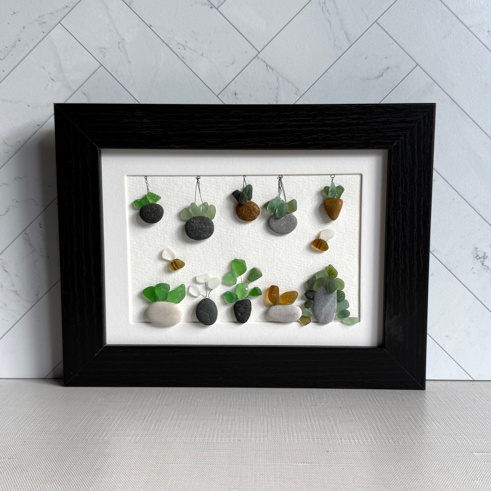 Framed artwork of small plants made from stones and sea glass on a white background