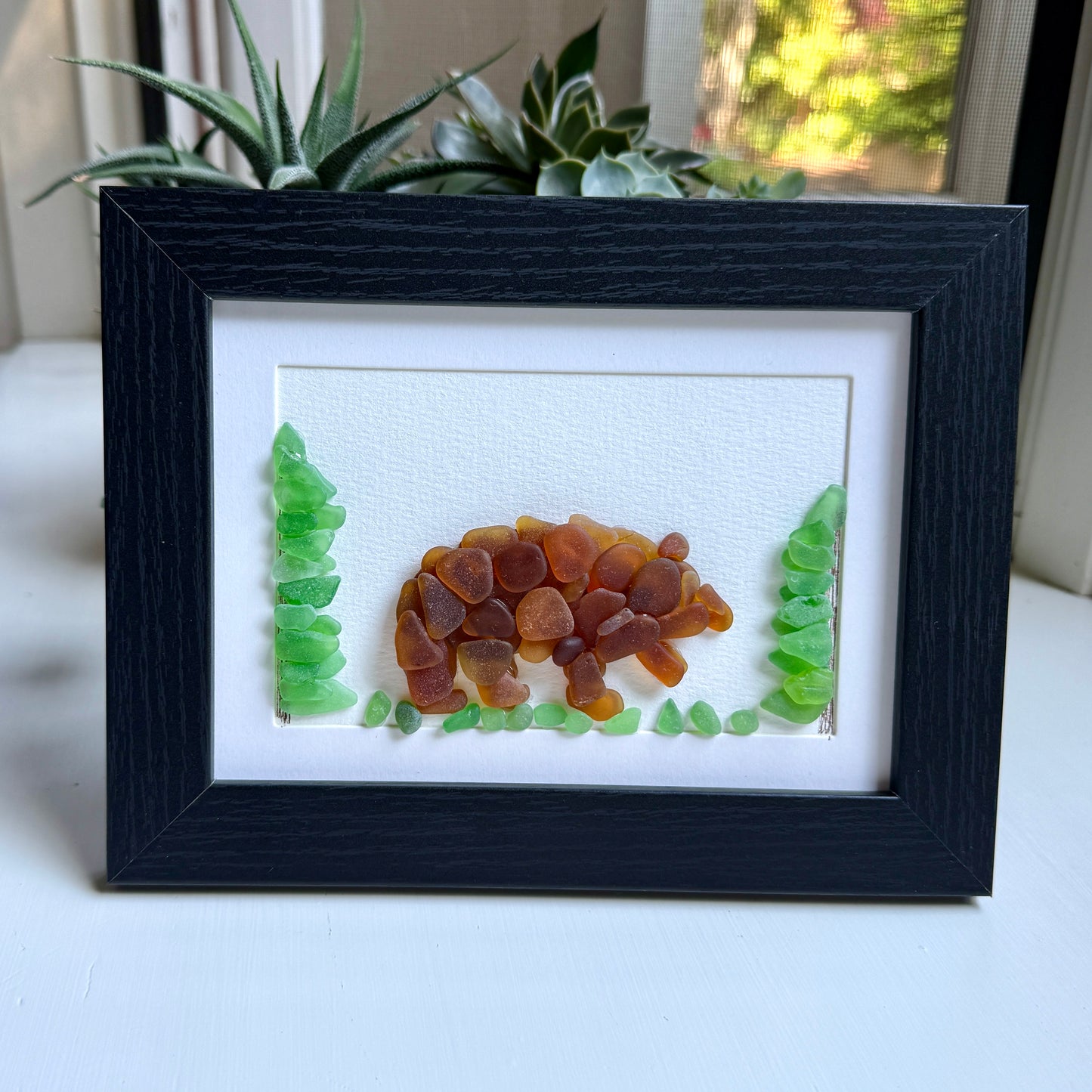 Framed artwork of a bear made from natural materials on a white surface with plants in the background.