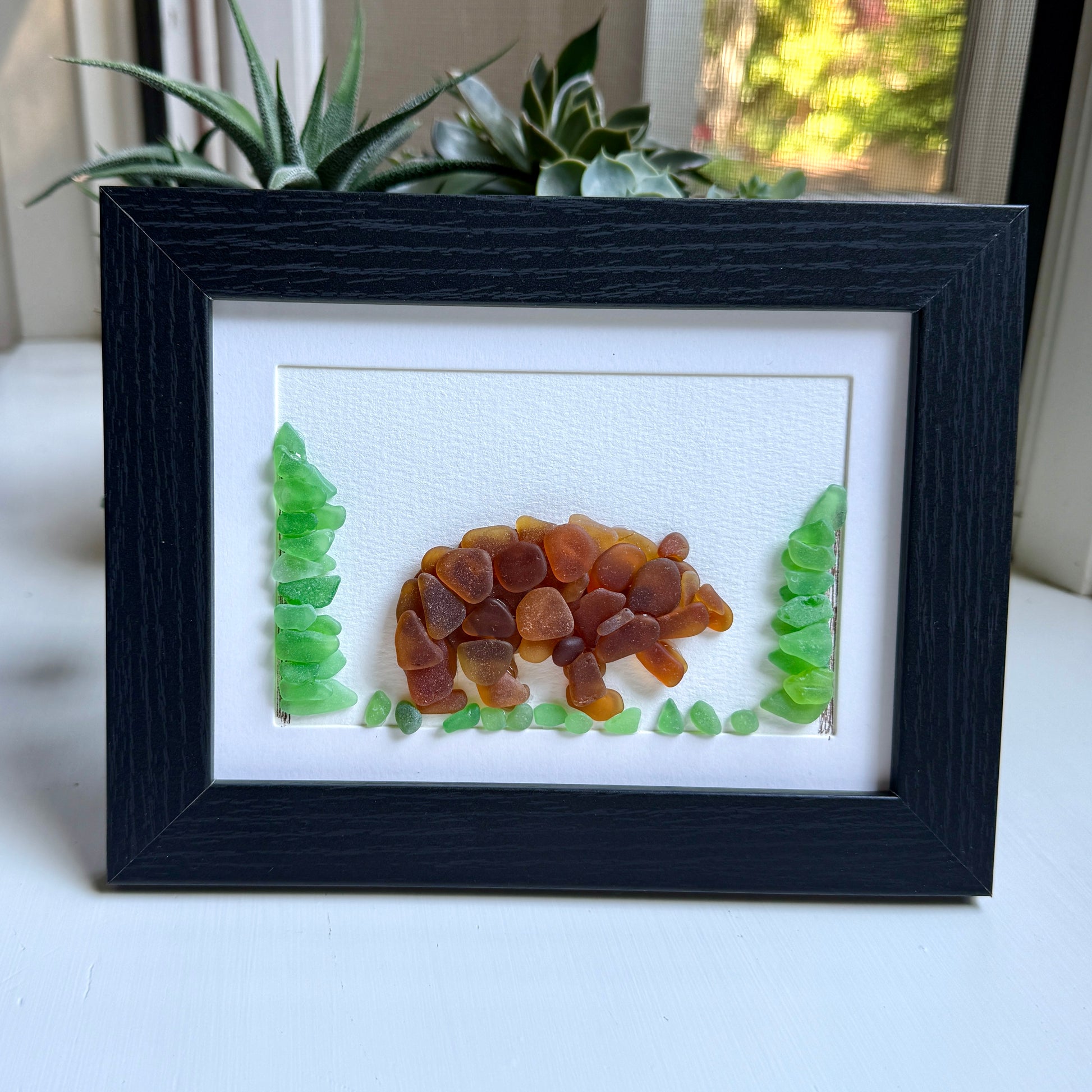 Framed artwork of a bear made from natural materials on a white surface with plants in the background.