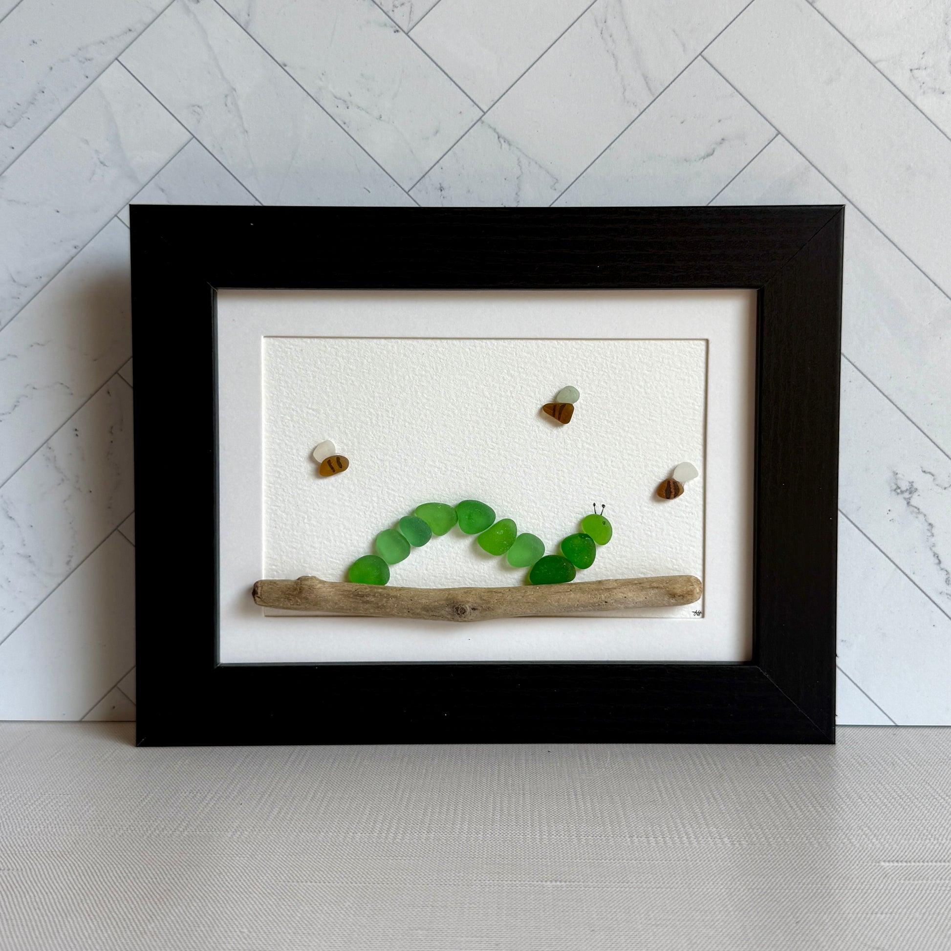 Framed artwork of a caterpillar made from green stones on a piece of wood against a marble wall.