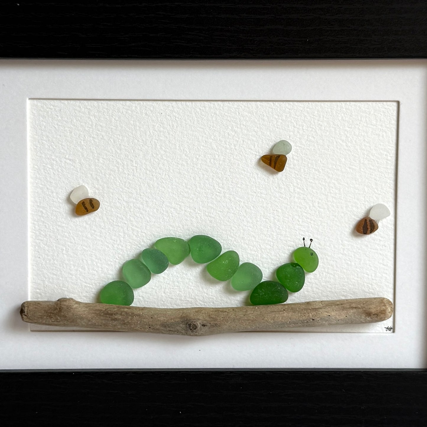 Artistic display of a caterpillar made from green glass pieces on a stick, framed in white.