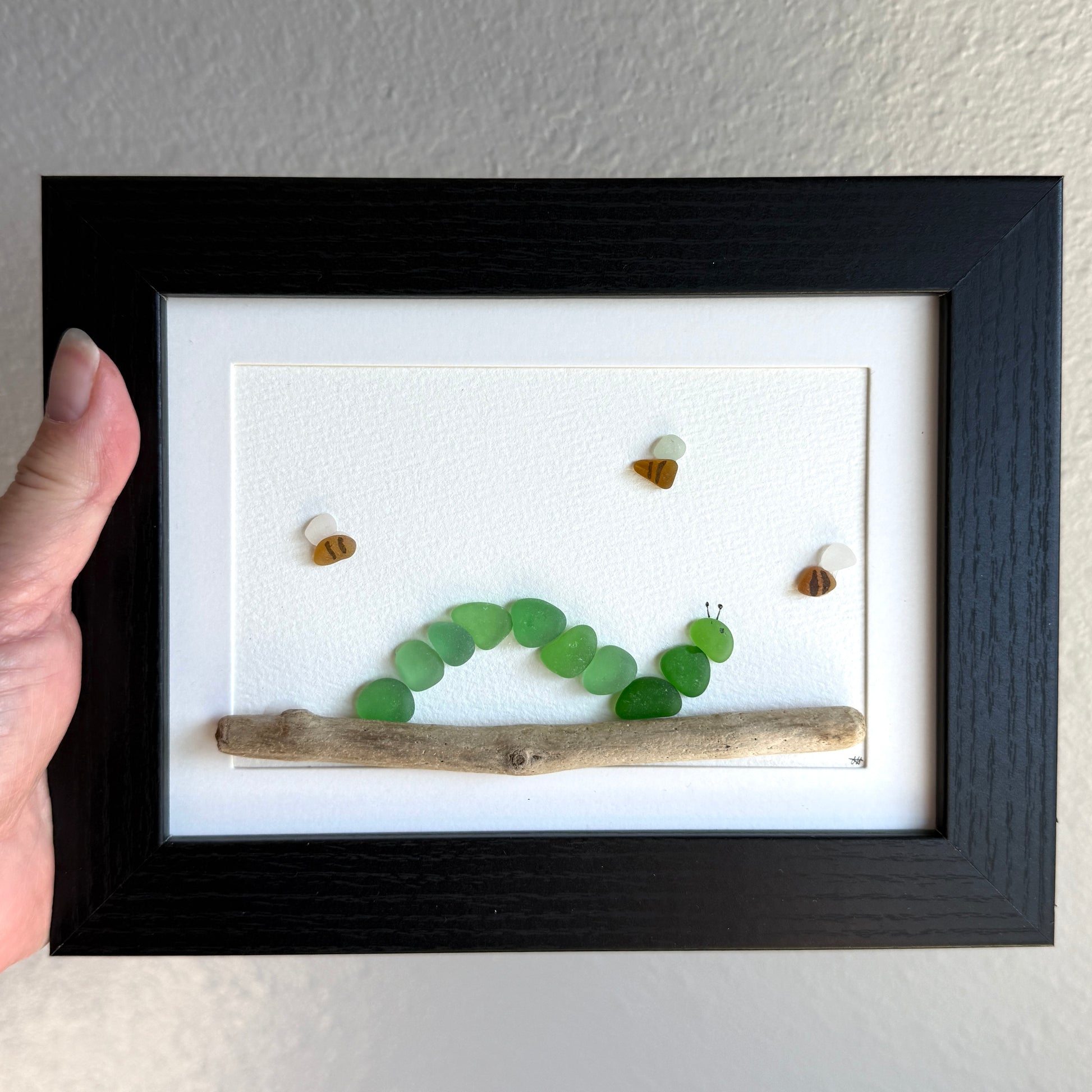 Framed artwork of a green caterpillar made from pebbles on a branch, held by a hand.