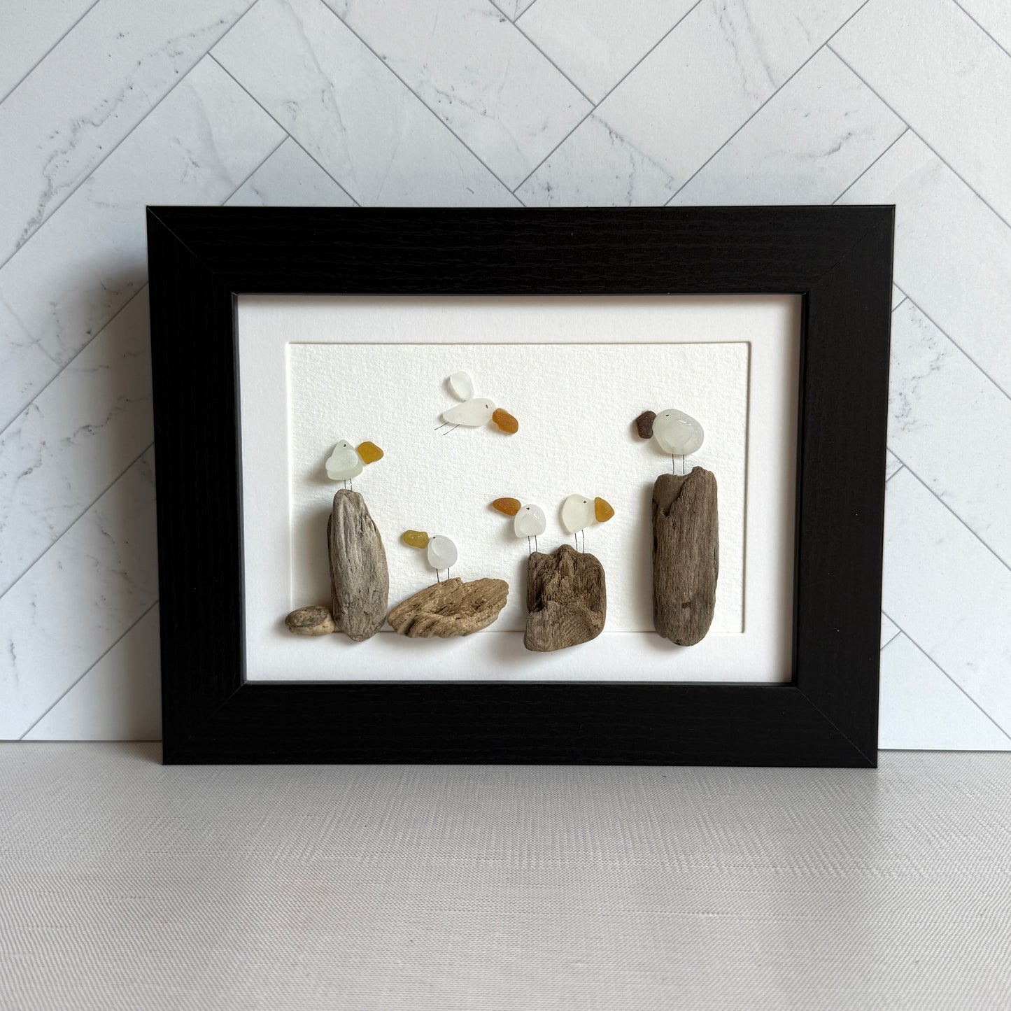 Framed sea glass artwork made of sea glass and driftwood on a white surface.