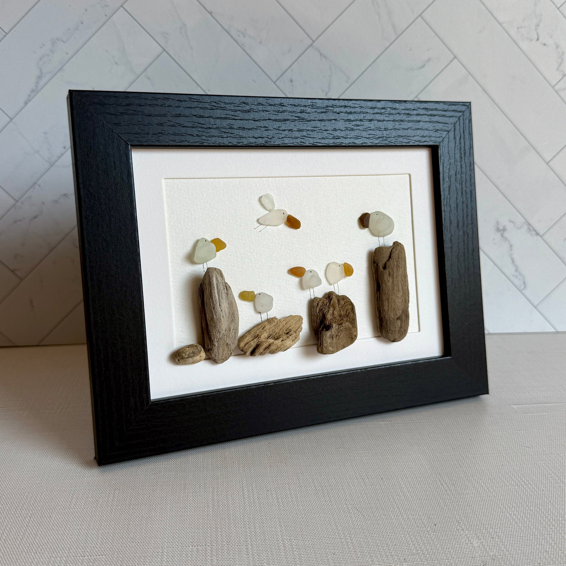 Framed sea glass artwork made of sea glass and driftwood on a white surface.