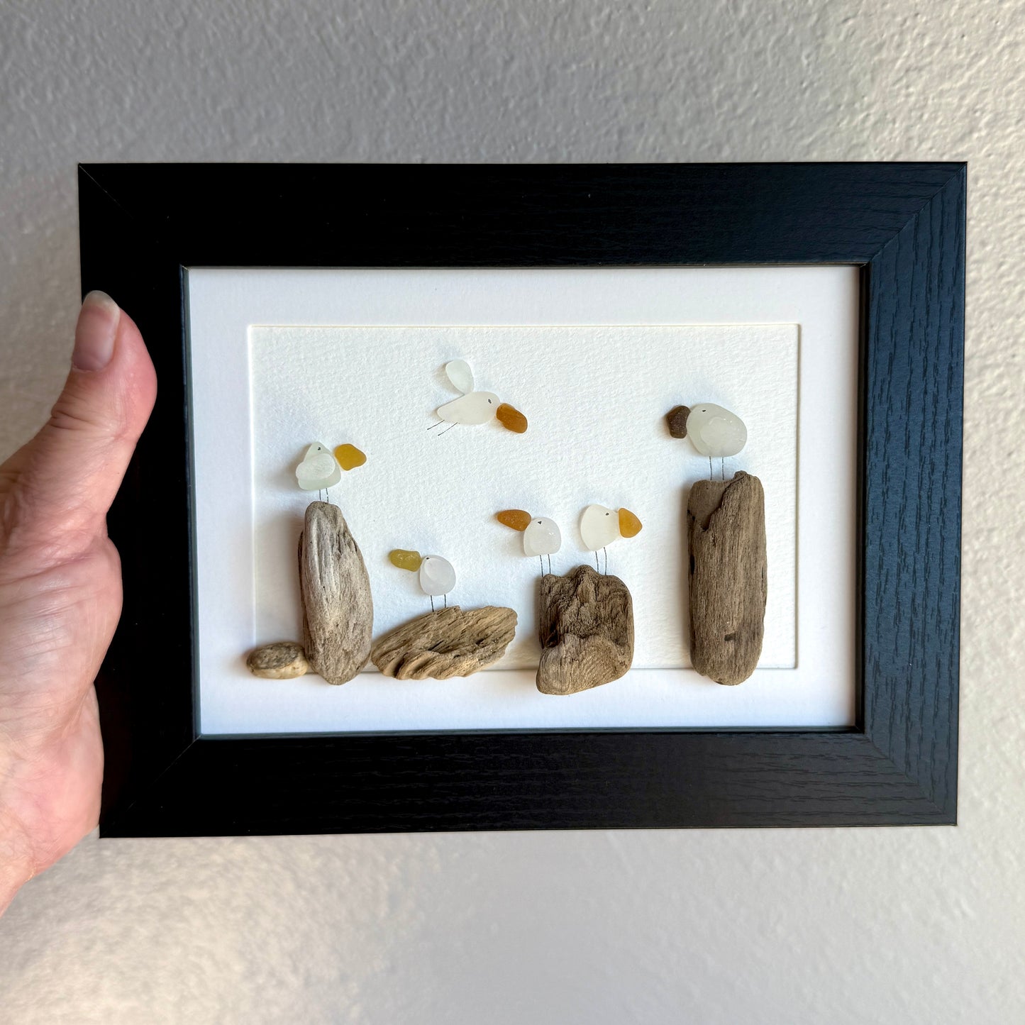 Framed sea glass artwork made of sea glass and driftwood on a white surface.