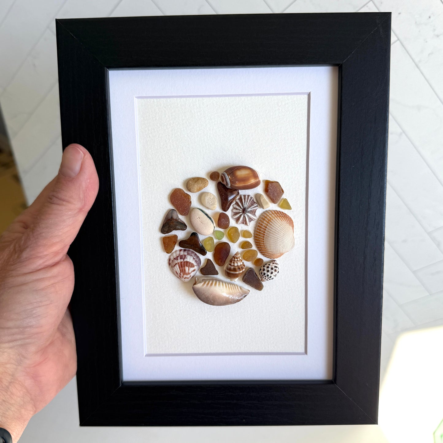 Framed artwork made of seashells held by a hand on a light background