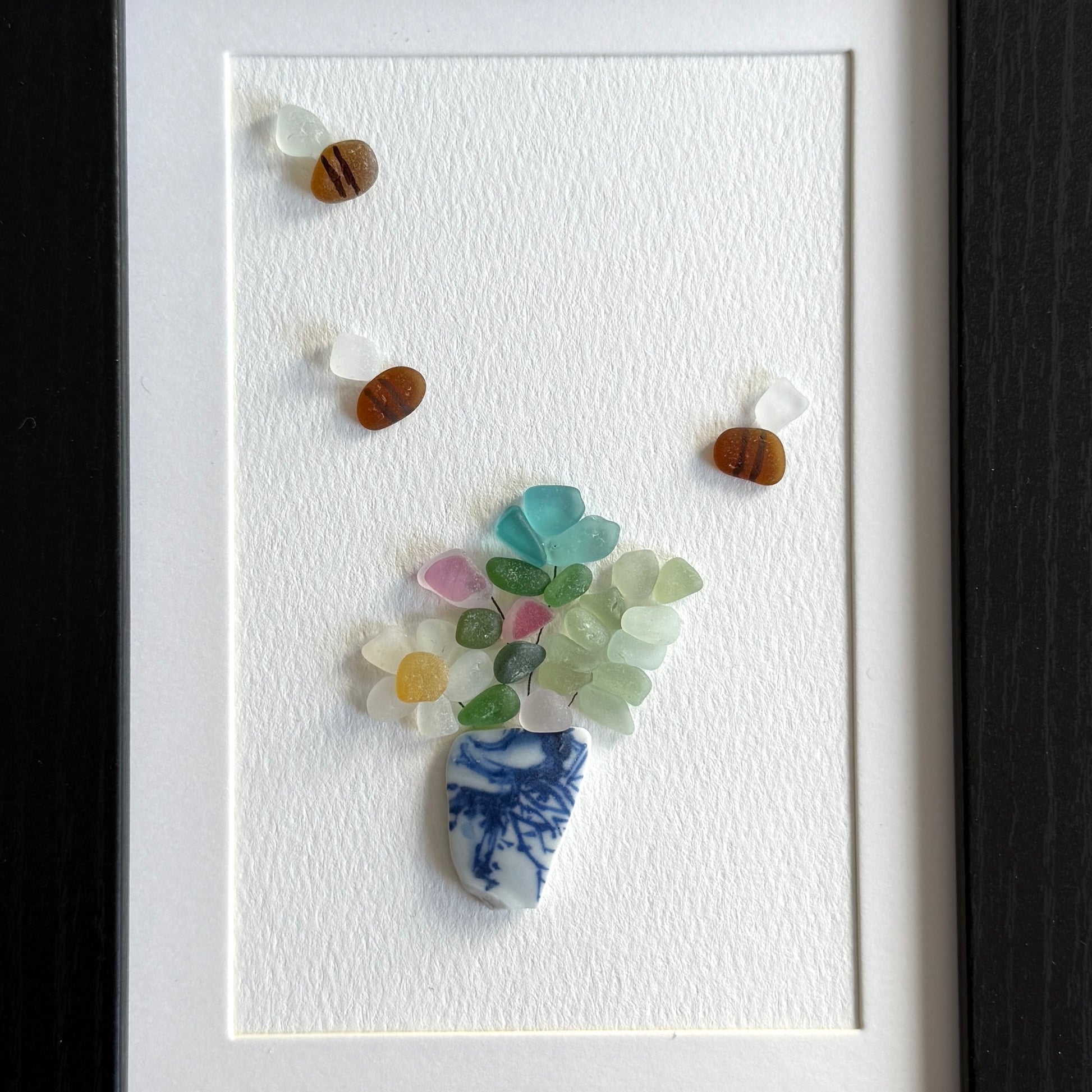 Framed artwork made from colorful sea glass shaped like a flower with a blue and white vase.