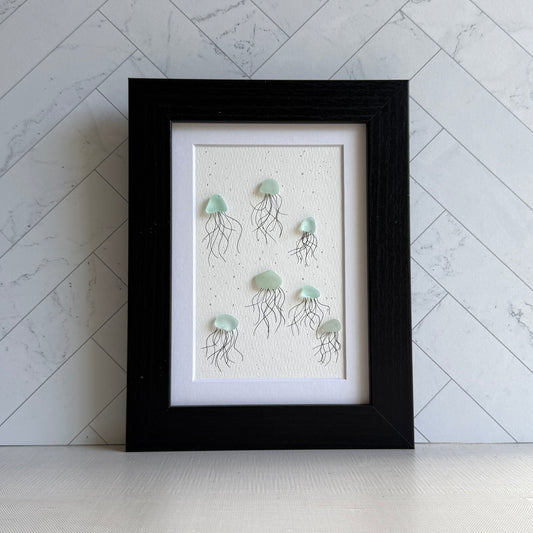 Framed sea glass artwork of jellyfish on a textured white background with a black frame.