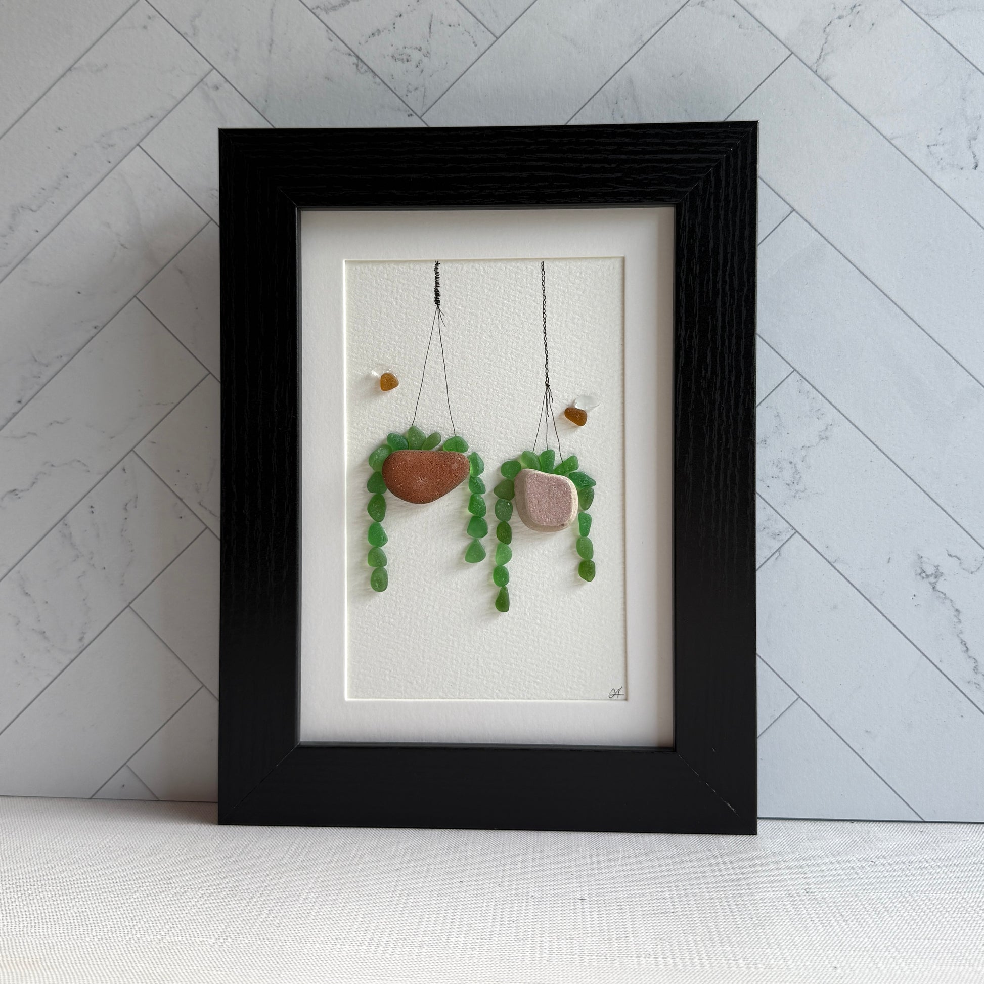 Framed sea glass artwork of hanging plant decorations on a white wall.