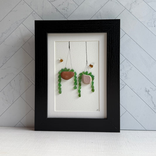 Framed sea glass artwork of hanging plant decorations on a white wall.