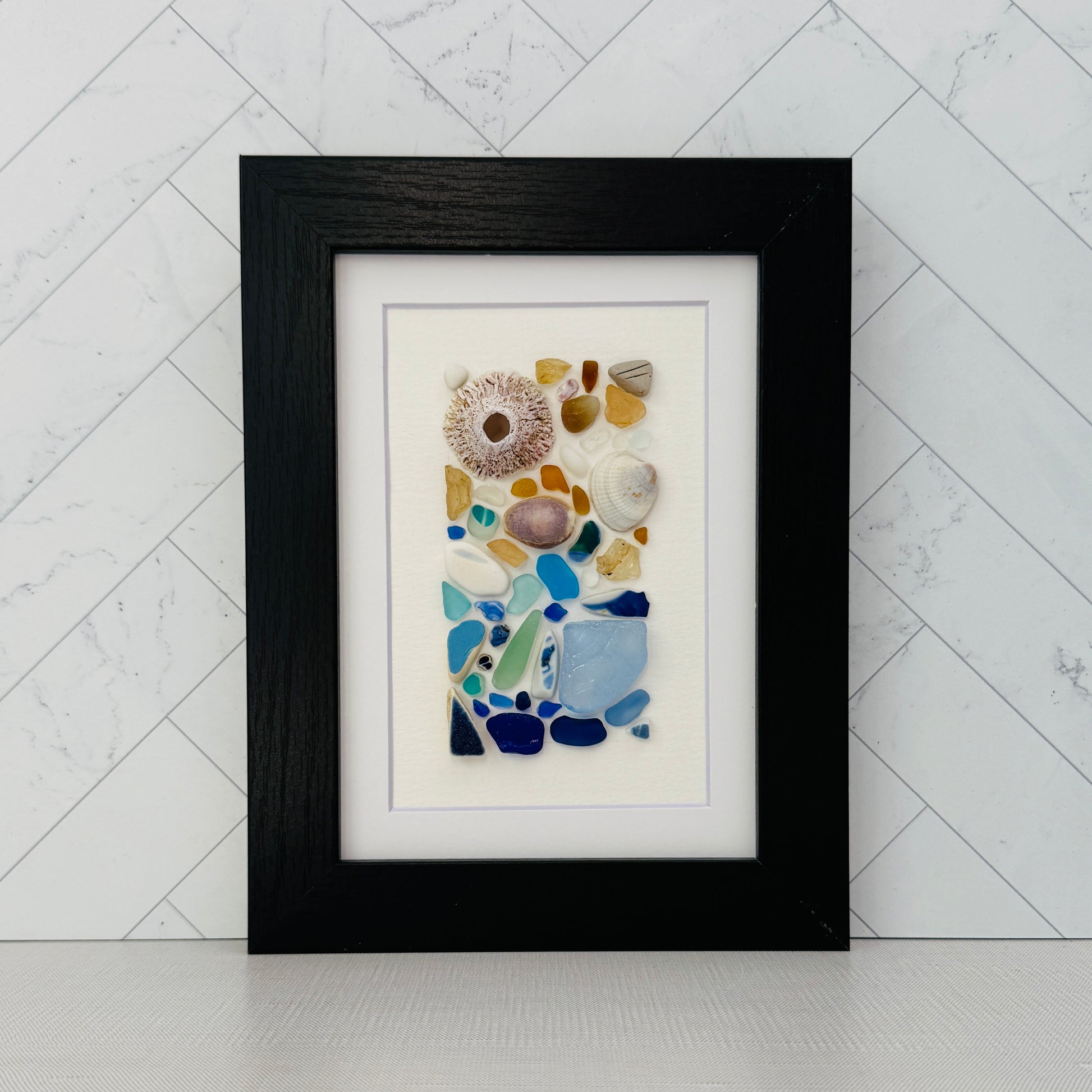 Handmade 5×7 framed sea-glass wall art featuring a mosaic of cobalt blue, aqua, white, and amber sea glass with seashells and coral pieces arranged vertically on white textured paper in a black wooden frame.