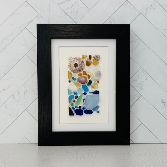 Handmade 5×7 framed sea-glass wall art featuring a mosaic of cobalt blue, aqua, white, and amber sea glass with seashells and coral pieces arranged vertically on white textured paper in a black wooden frame.