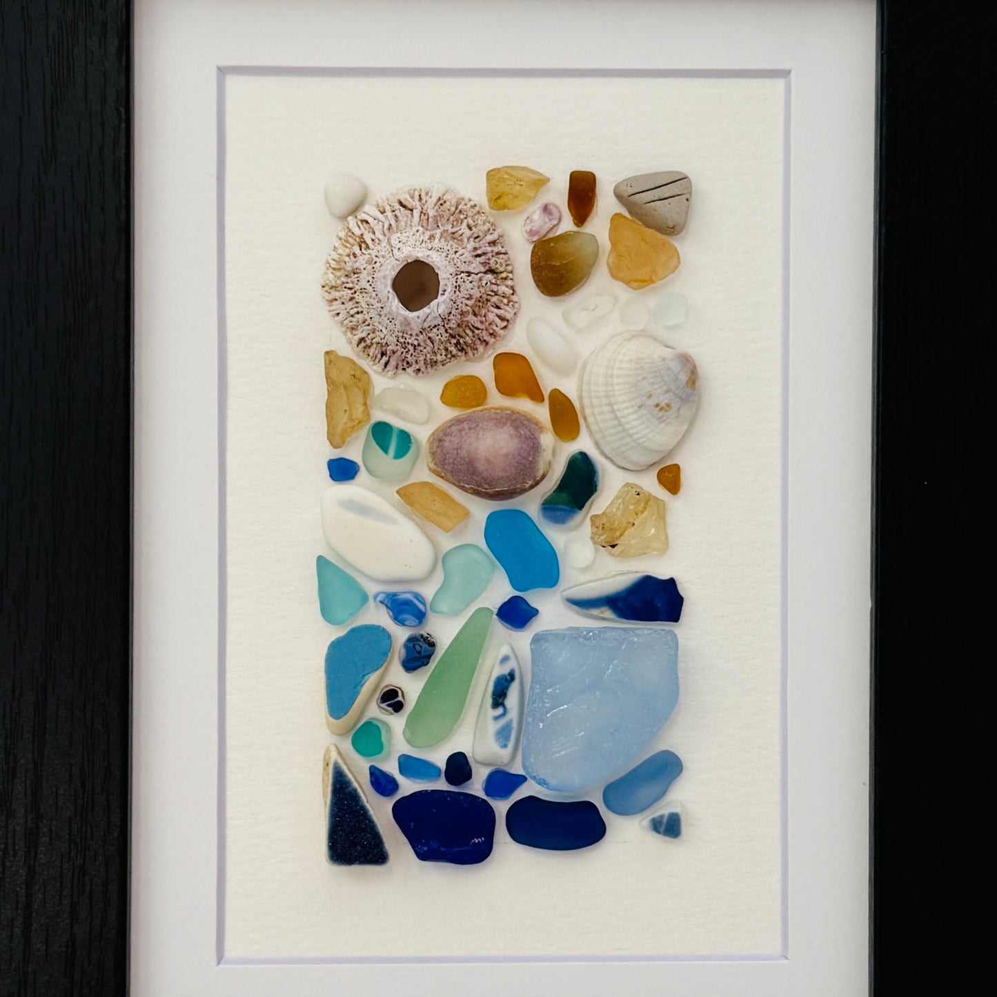 Handmade 5×7 framed sea-glass wall art featuring a mosaic of cobalt blue, aqua, white, and amber sea glass with seashells and coral pieces arranged vertically on white textured paper in a black wooden frame.