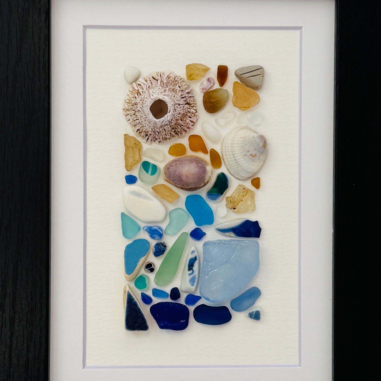 Handmade 5×7 framed sea-glass wall art featuring a mosaic of cobalt blue, aqua, white, and amber sea glass with seashells and coral pieces arranged vertically on white textured paper in a black wooden frame.