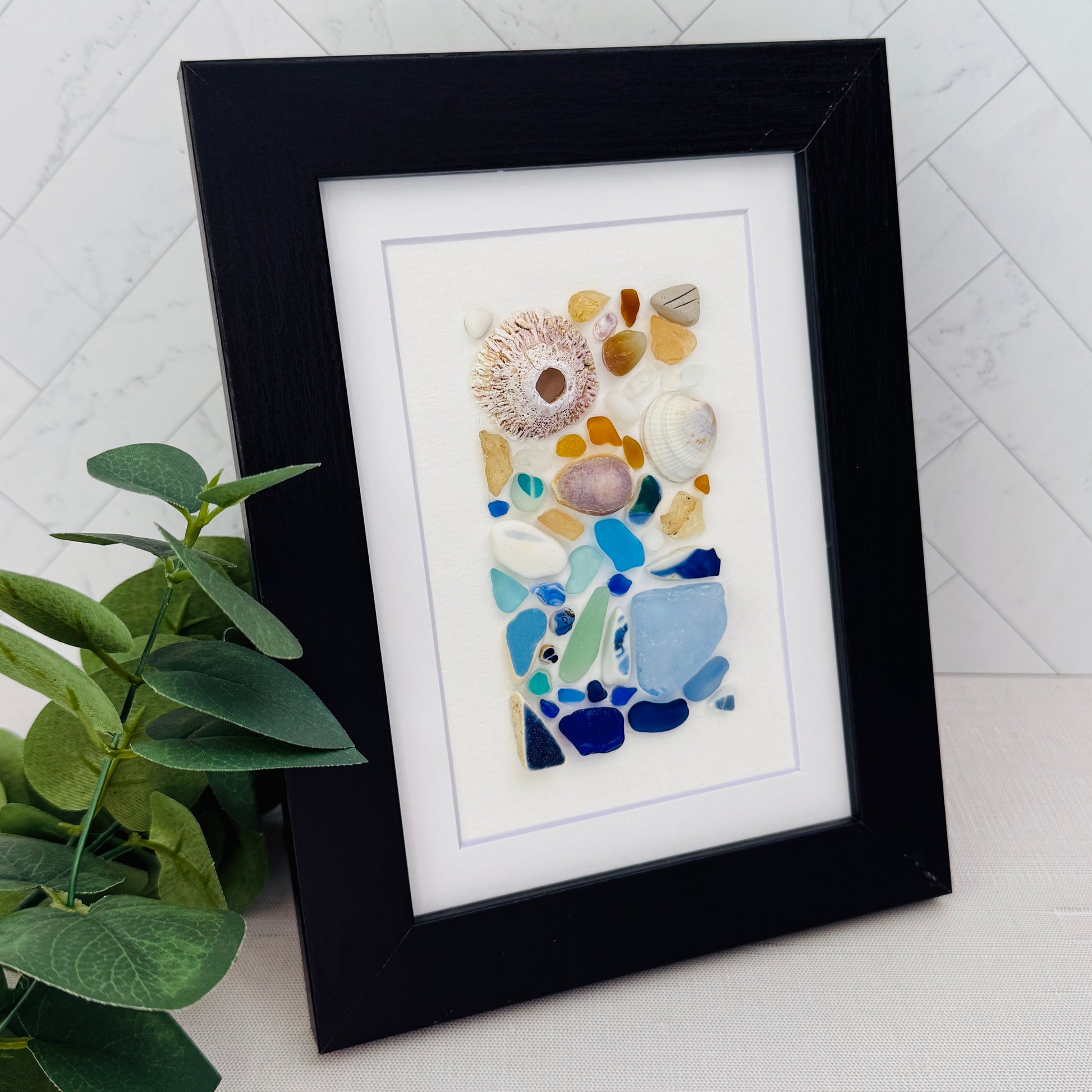 Handmade 5×7 framed sea-glass wall art featuring a mosaic of cobalt blue, aqua, white, and amber sea glass with seashells and coral pieces arranged vertically on white textured paper in a black wooden frame.