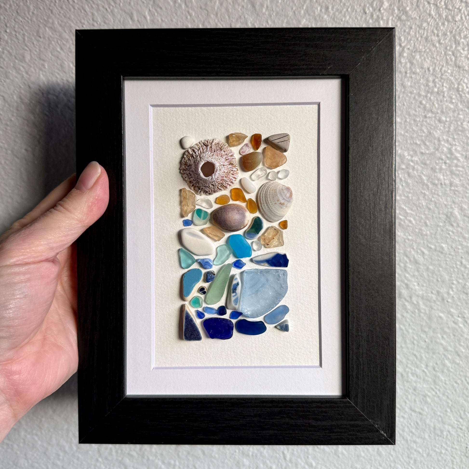 Handmade 5×7 framed sea-glass wall art featuring a mosaic of cobalt blue, aqua, white, and amber sea glass with seashells and coral pieces arranged vertically on white textured paper in a black wooden frame.
