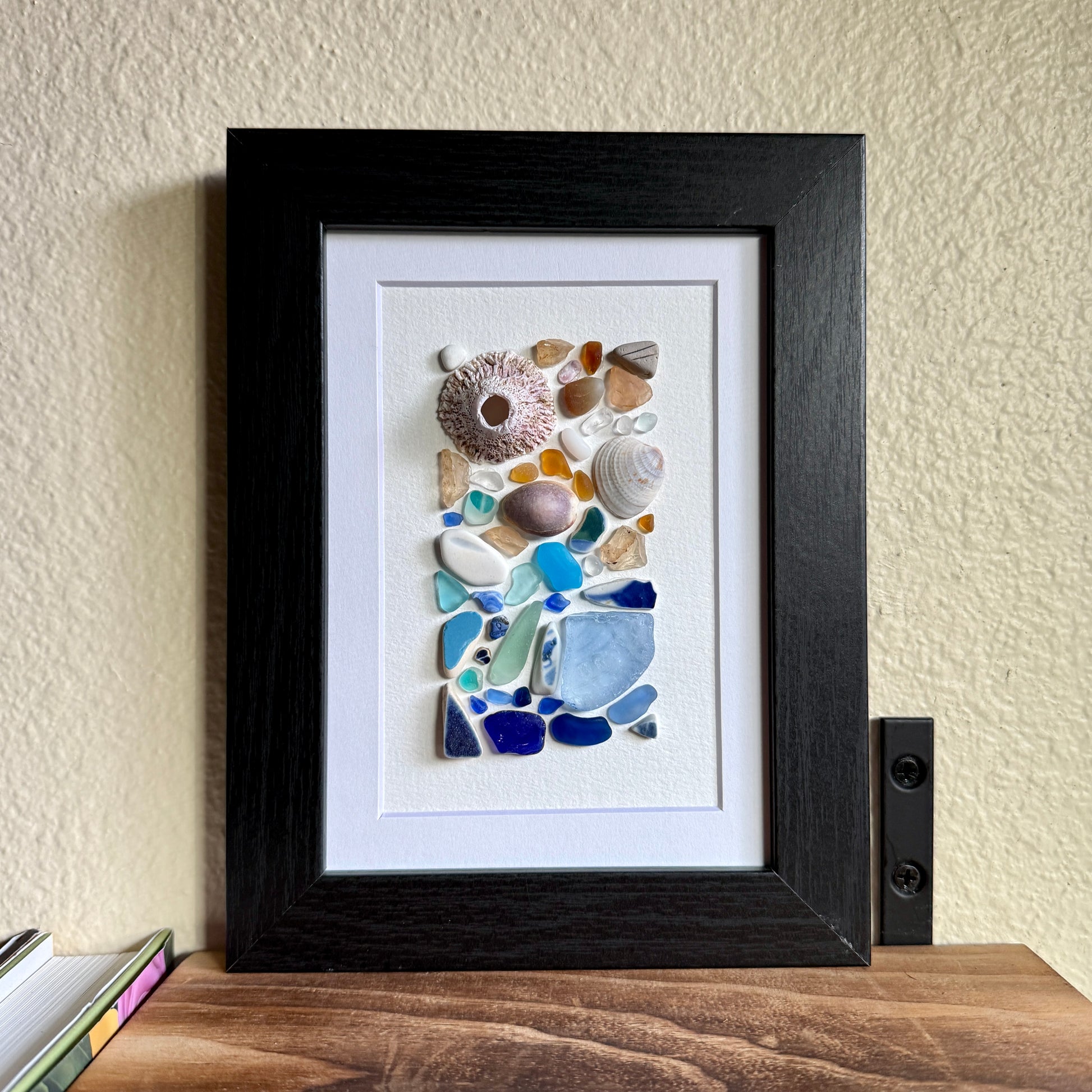 Handmade 5×7 framed sea-glass wall art featuring a mosaic of cobalt blue, aqua, white, and amber sea glass with seashells and coral pieces arranged vertically on white textured paper in a black wooden frame.