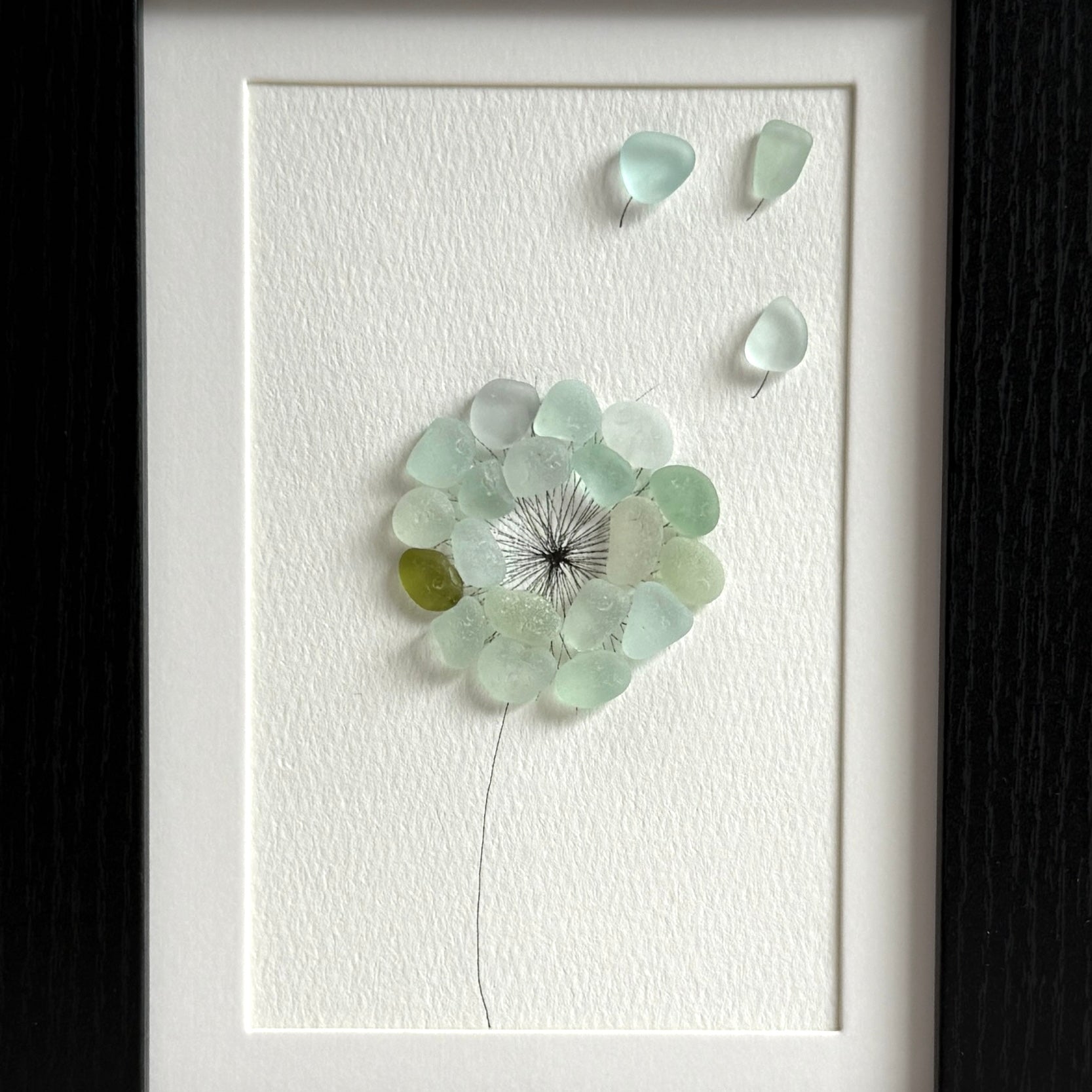 This is a framed 5 x 7 inch sea glass art picture featuring a single aqua blue and sea foam dandelion with a couple fly-aways.  This item is perfect for gift giving and ready to hang or place on a shelf.
