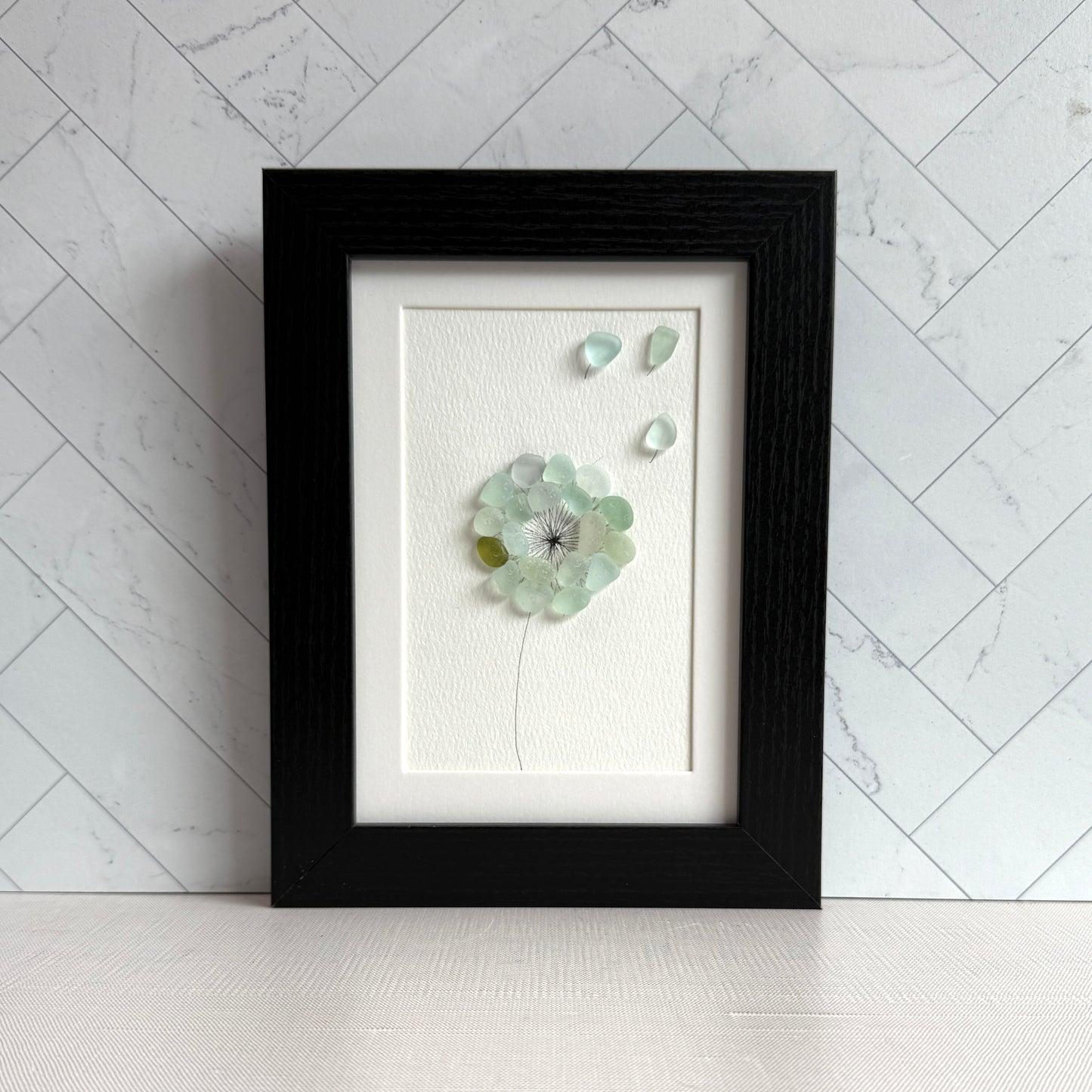This is a framed 5 x 7 inch sea glass art picture featuring a single aqua blue and sea foam dandelion with a couple fly-aways.  This item is perfect for gift giving and ready to hang or place on a shelf.