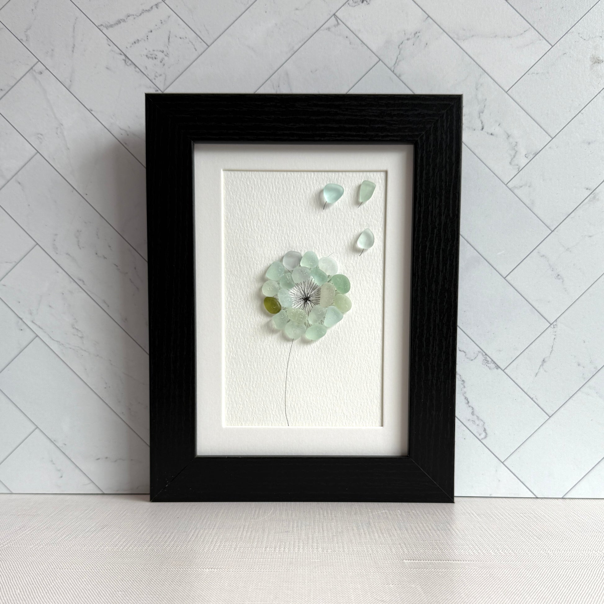 This is a framed 5 x 7 inch sea glass art picture featuring a single aqua blue and sea foam dandelion with a couple fly-aways.  This item is perfect for gift giving and ready to hang or place on a shelf.