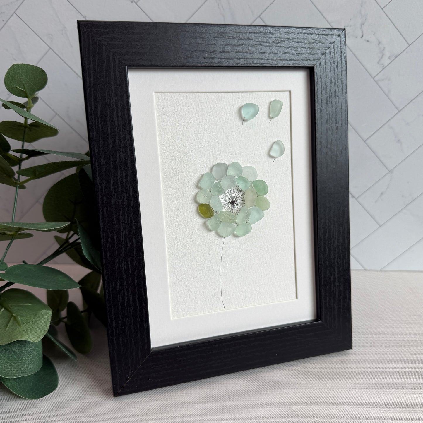This is a framed 5 x 7 inch sea glass art picture featuring a single aqua blue and sea foam dandelion with a couple fly-aways.  This item is perfect for gift giving and ready to hang or place on a shelf.