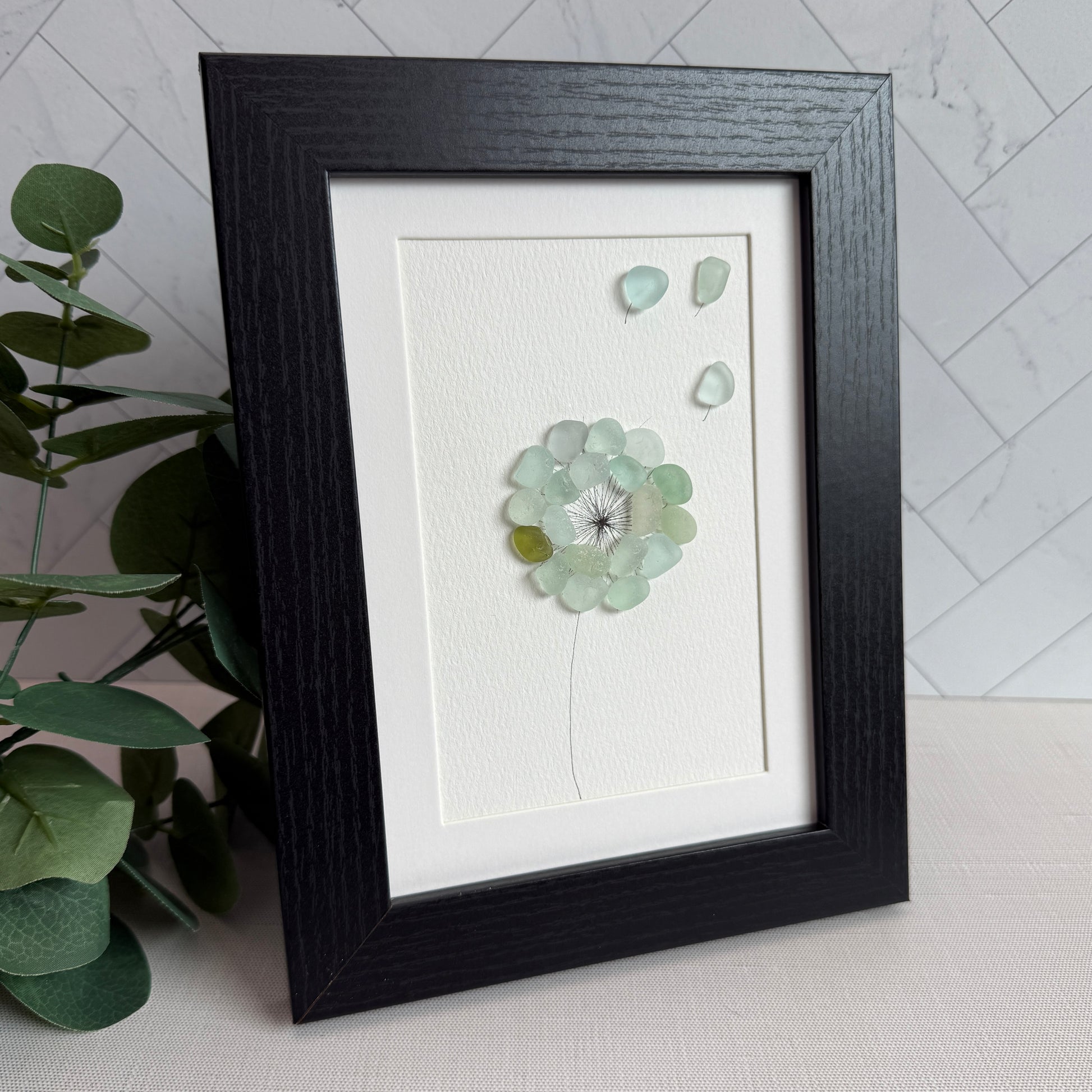 This is a framed 5 x 7 inch sea glass art picture featuring a single aqua blue and sea foam dandelion with a couple fly-aways.  This item is perfect for gift giving and ready to hang or place on a shelf.
