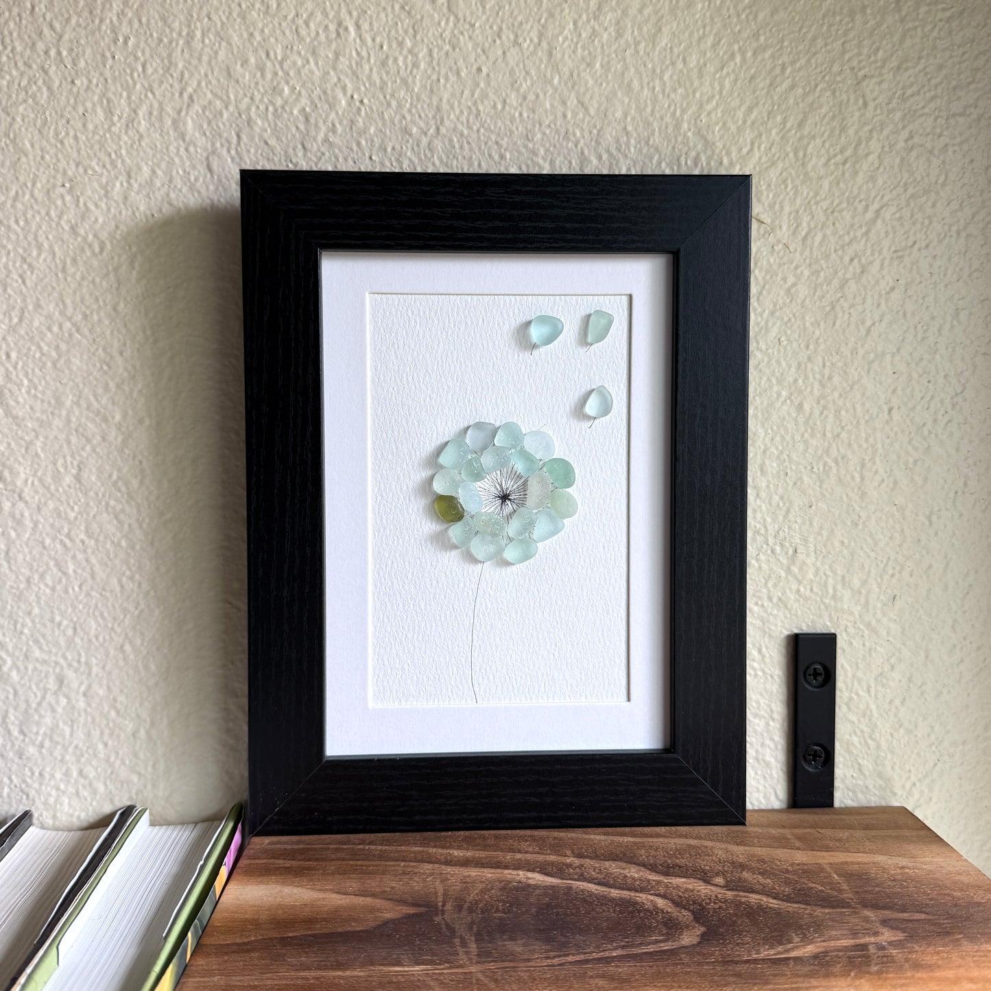 This is a framed 5 x 7 inch sea glass art picture featuring a single aqua blue and sea foam dandelion with a couple fly-aways.  This item is perfect for gift giving and ready to hang or place on a shelf.