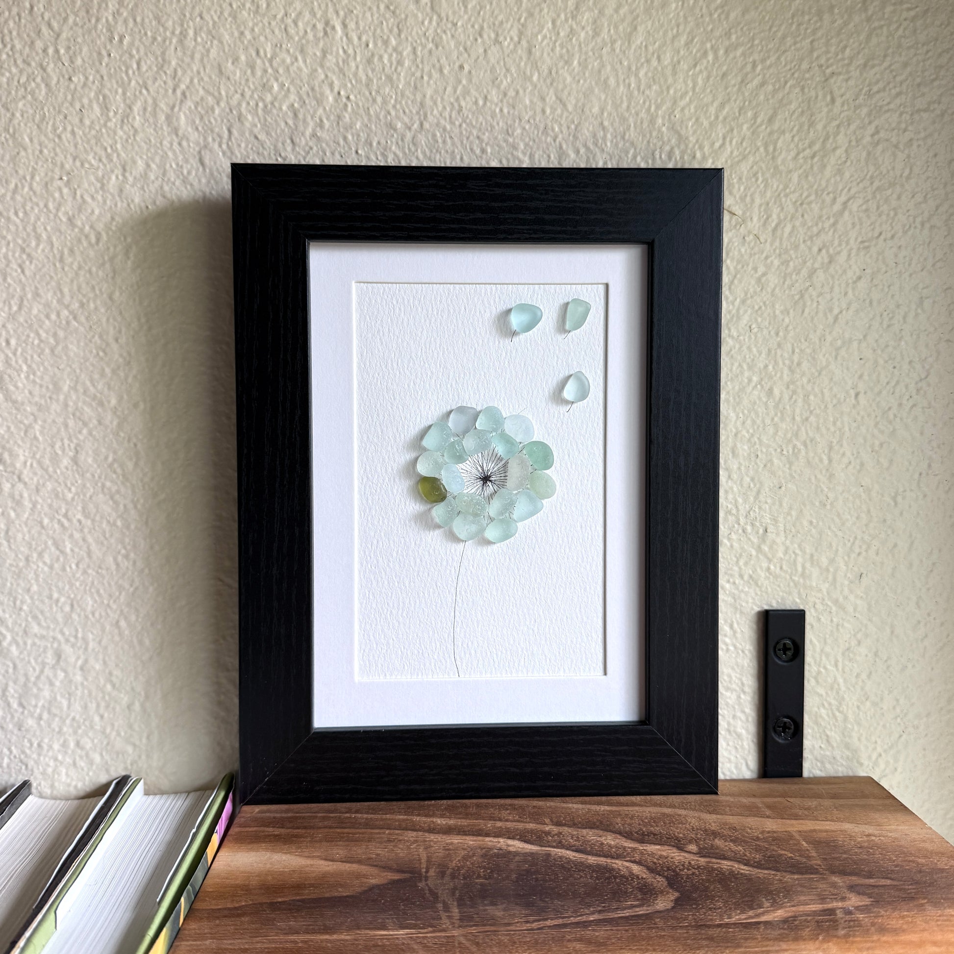 This is a framed 5 x 7 inch sea glass art picture featuring a single aqua blue and sea foam dandelion with a couple fly-aways.  This item is perfect for gift giving and ready to hang or place on a shelf.