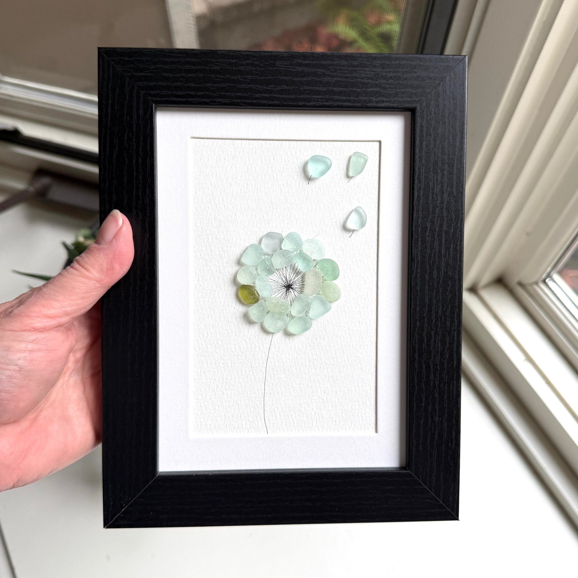 This is a framed 5 x 7 inch sea glass art picture featuring a single aqua blue and sea foam dandelion with a couple fly-aways.  This item is perfect for gift giving and ready to hang or place on a shelf.