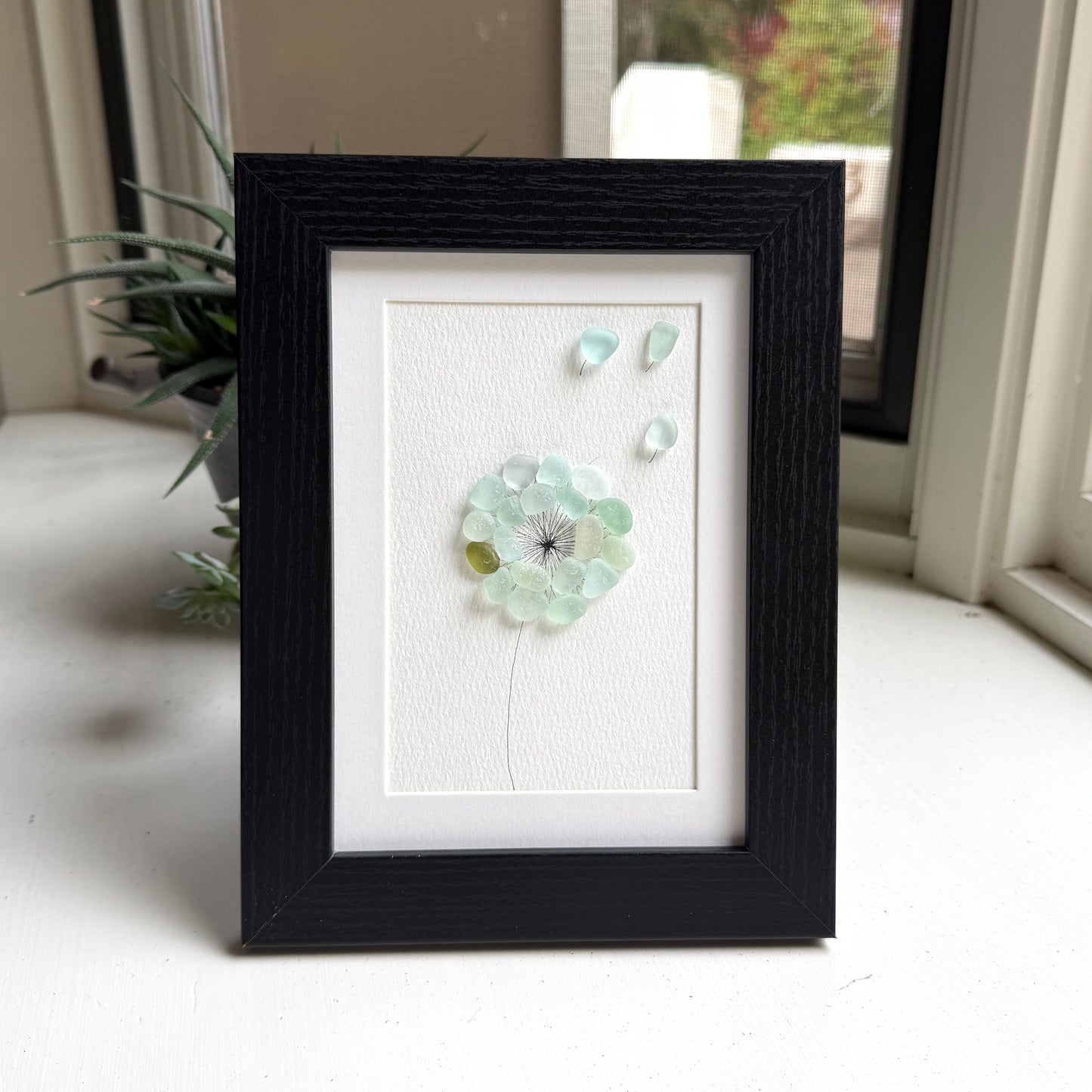 This is a framed 5 x 7 inch sea glass art picture featuring a single aqua blue and sea foam dandelion with a couple fly-aways.  This item is perfect for gift giving and ready to hang or place on a shelf.