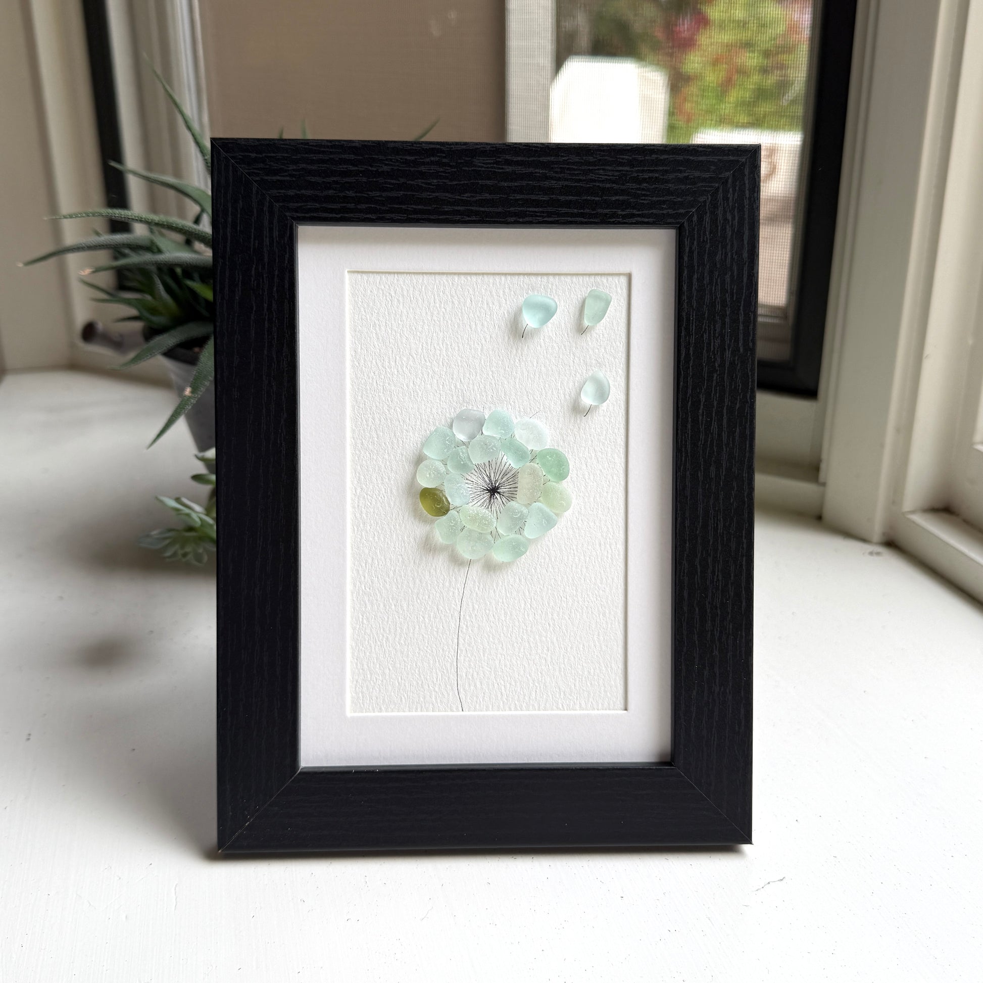 This is a framed 5 x 7 inch sea glass art picture featuring a single aqua blue and sea foam dandelion with a couple fly-aways.  This item is perfect for gift giving and ready to hang or place on a shelf.