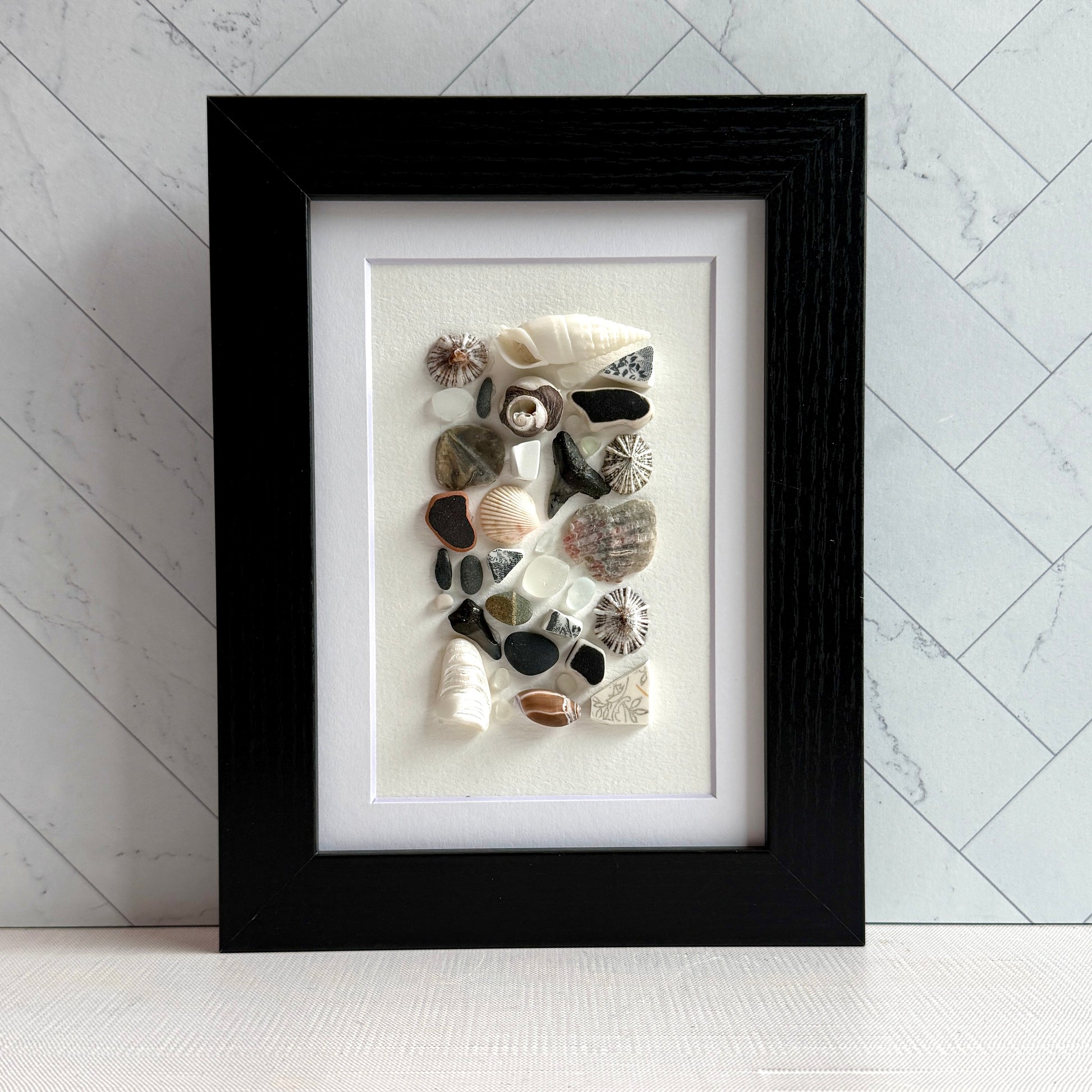 A 5×7 framed wall art piece featuring hand-arranged black, gray and white sea glass and beach-found shells and shark-tooth, in a sleek black wood frame, set against a white background.