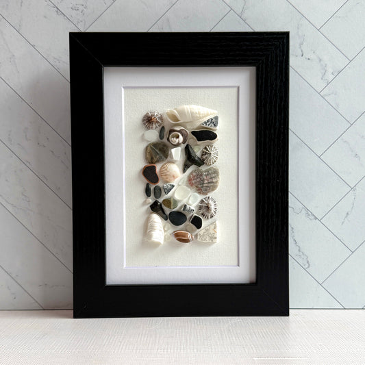 A 5×7 framed wall art piece featuring hand-arranged black, gray and white sea glass and beach-found shells and shark-tooth, in a sleek black wood frame, set against a white background.